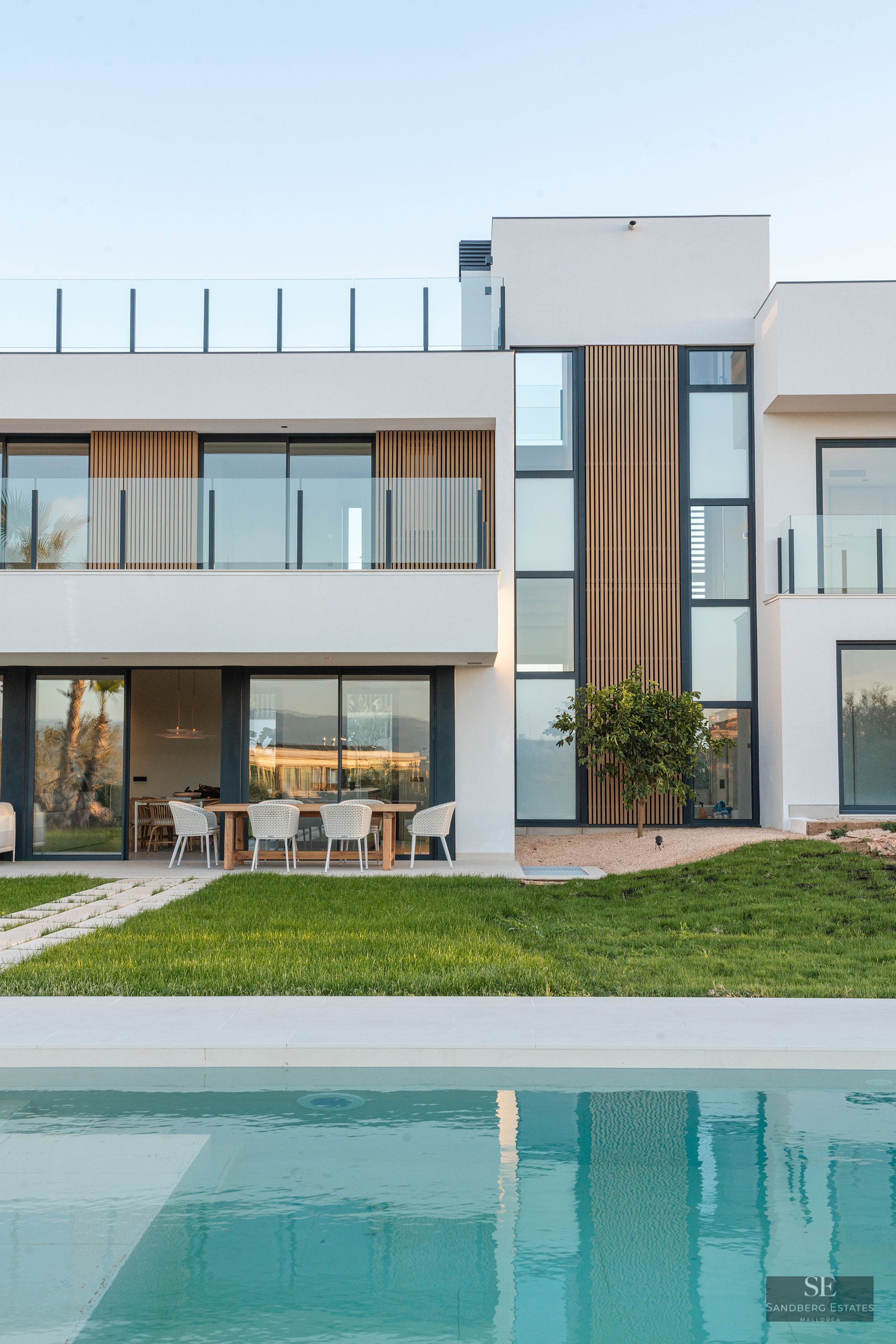 Modern white villa with wood accents, large windows, and a turquoise swimming pool in front of a green lawn.