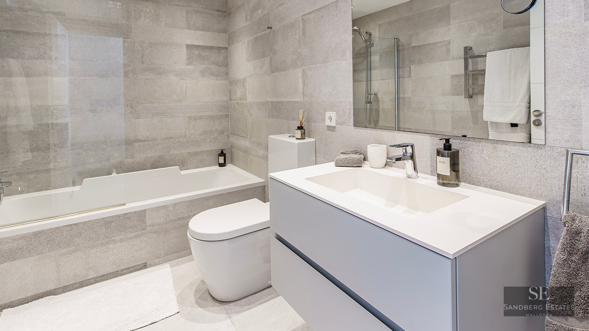 Contemporary bathroom featuring grey stone-effect tiles, a white bathtub, toilet, and modern vanity with a large mirror.