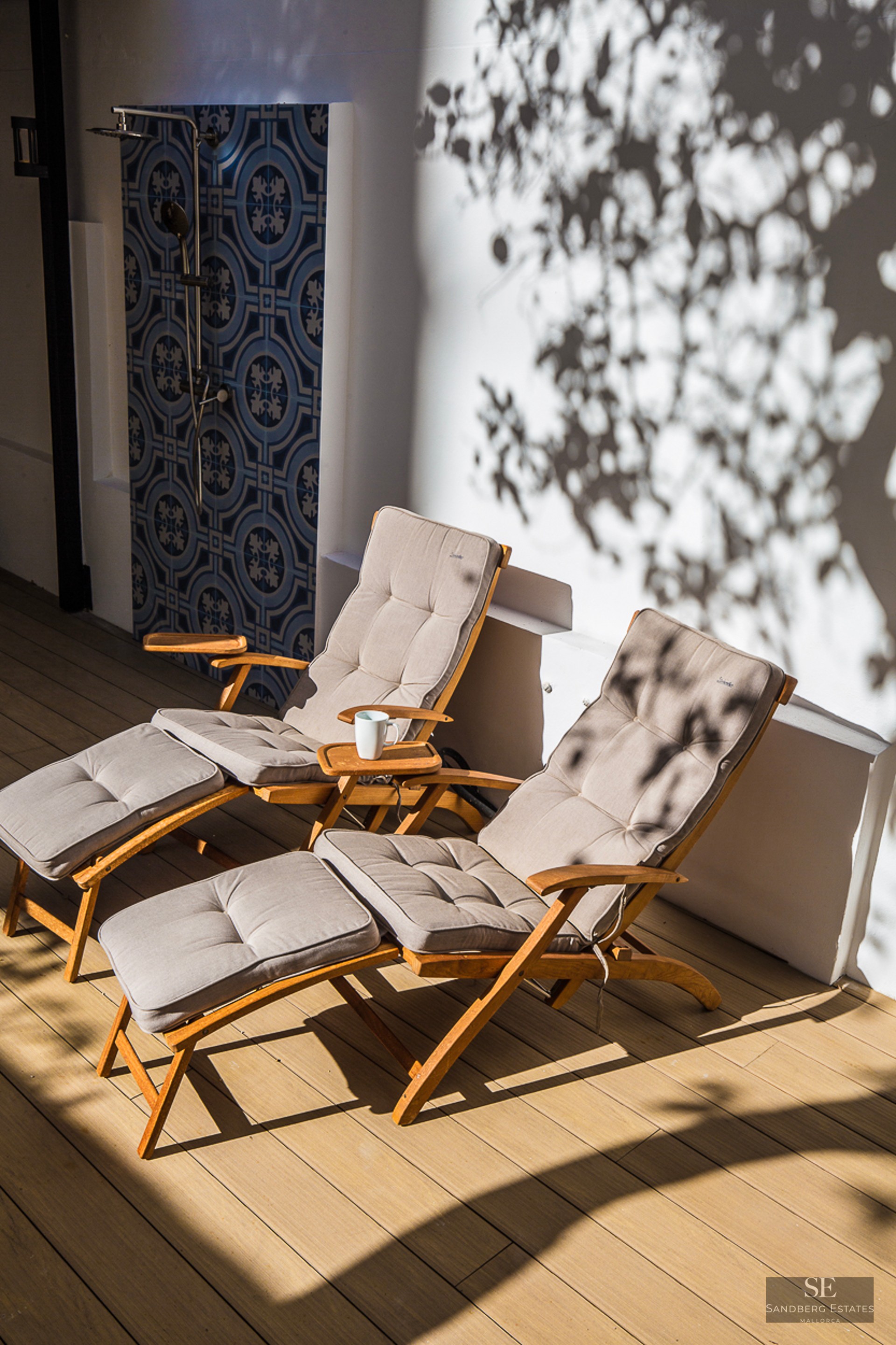 Two wooden lounge chairs on a deck next to a decorative blue tiled outdoor shower with leaf shadows on the wall.
