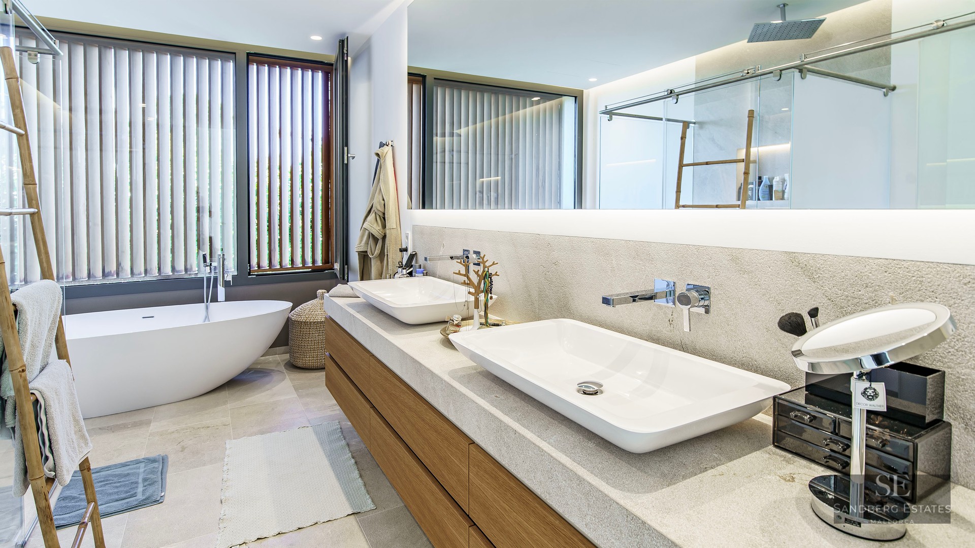 Salle de bain moderne avec double vasque, grand miroir éclairé et baignoire autoportante près d'une fenêtre à persiennes.