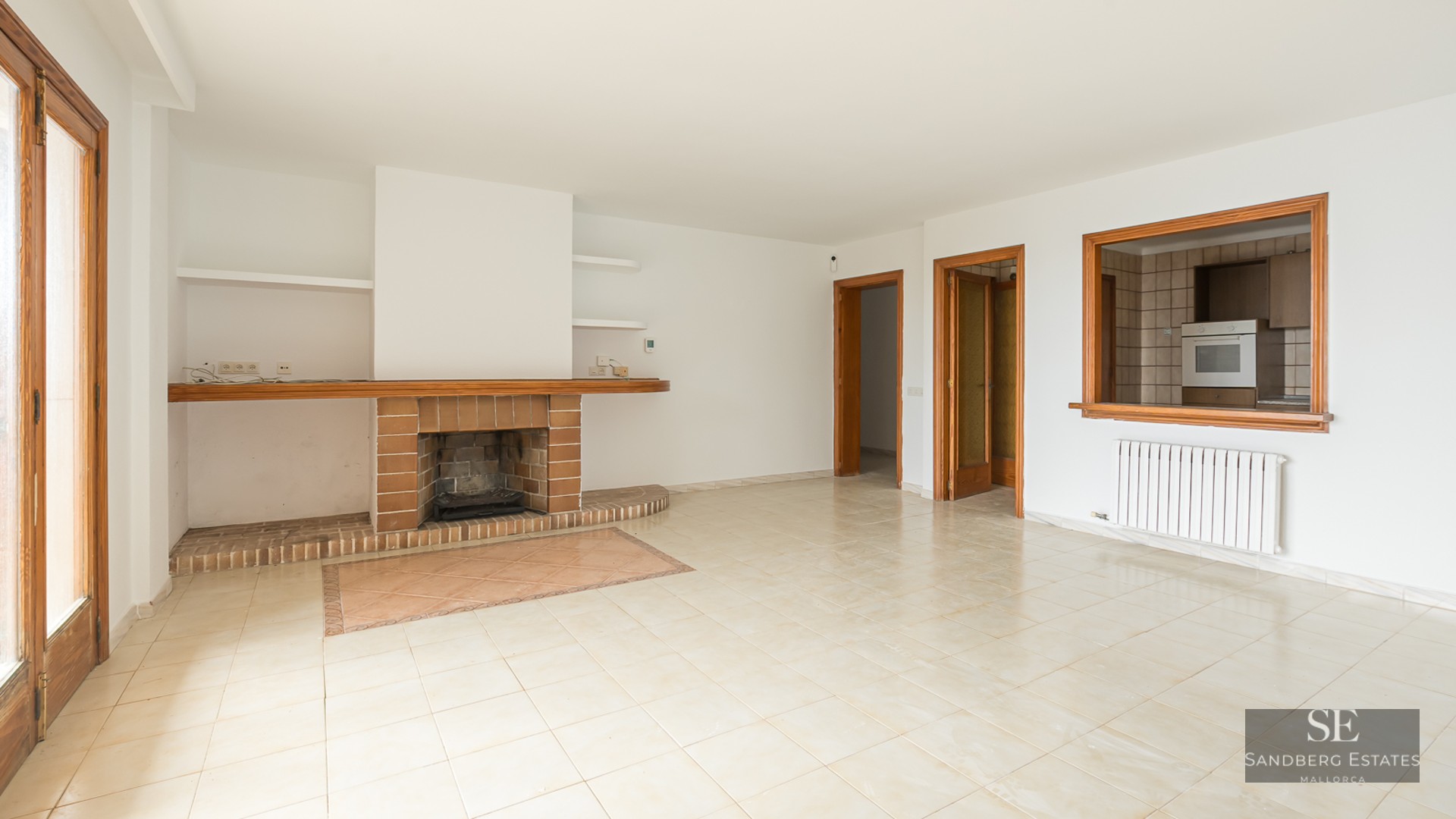 Empty living room with beige floor tiles, white walls, a brick fireplace, and a kitchen pass-through window.