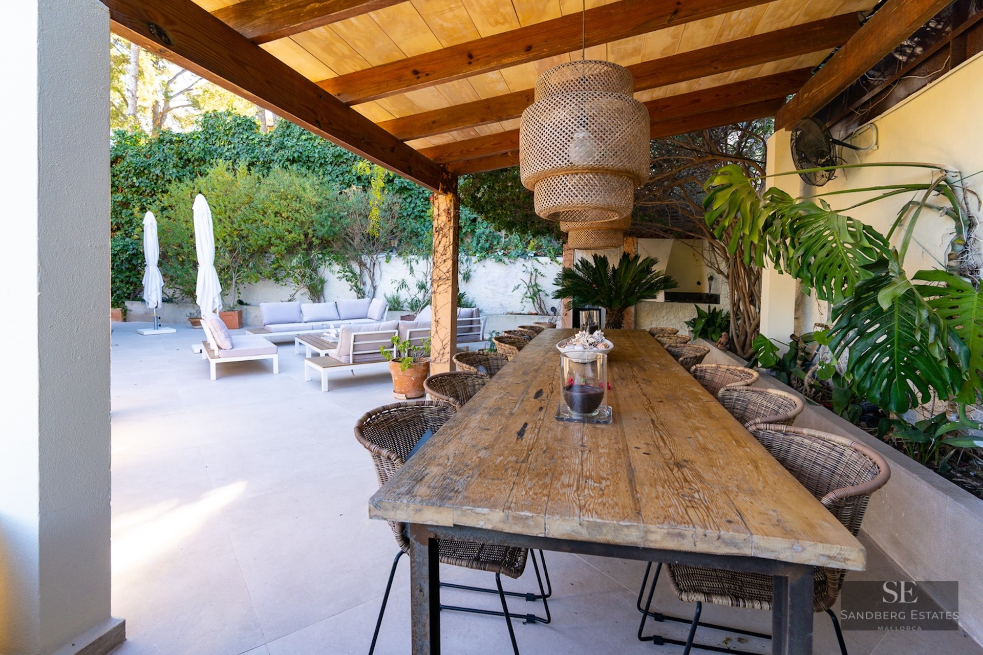 Long wooden dining table under a pergola with wicker chairs and a pendant light next to an outdoor lounge.