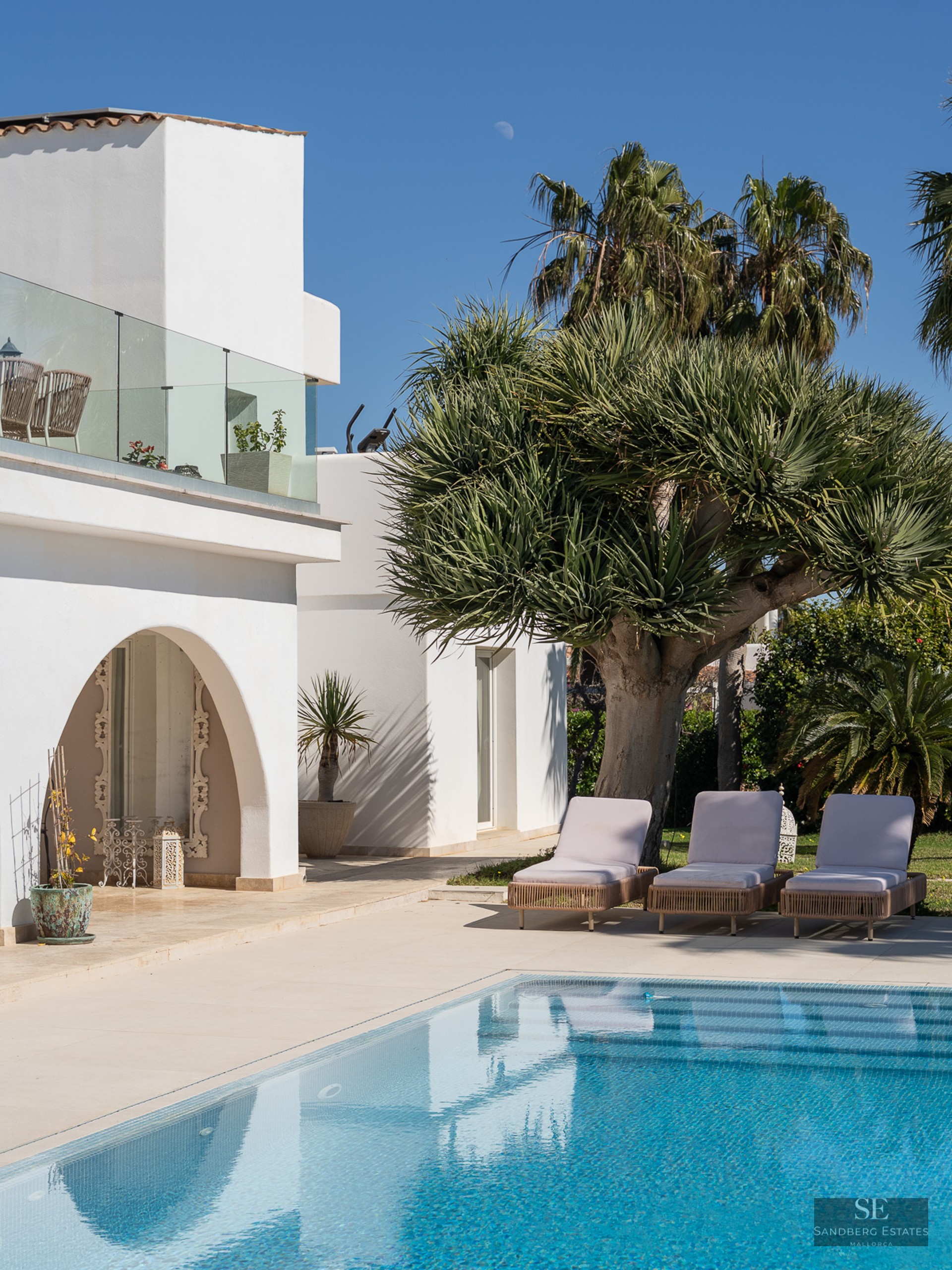 A bright swimming pool next to a white villa with three sun loungers and tropical palm trees under a blue sky.