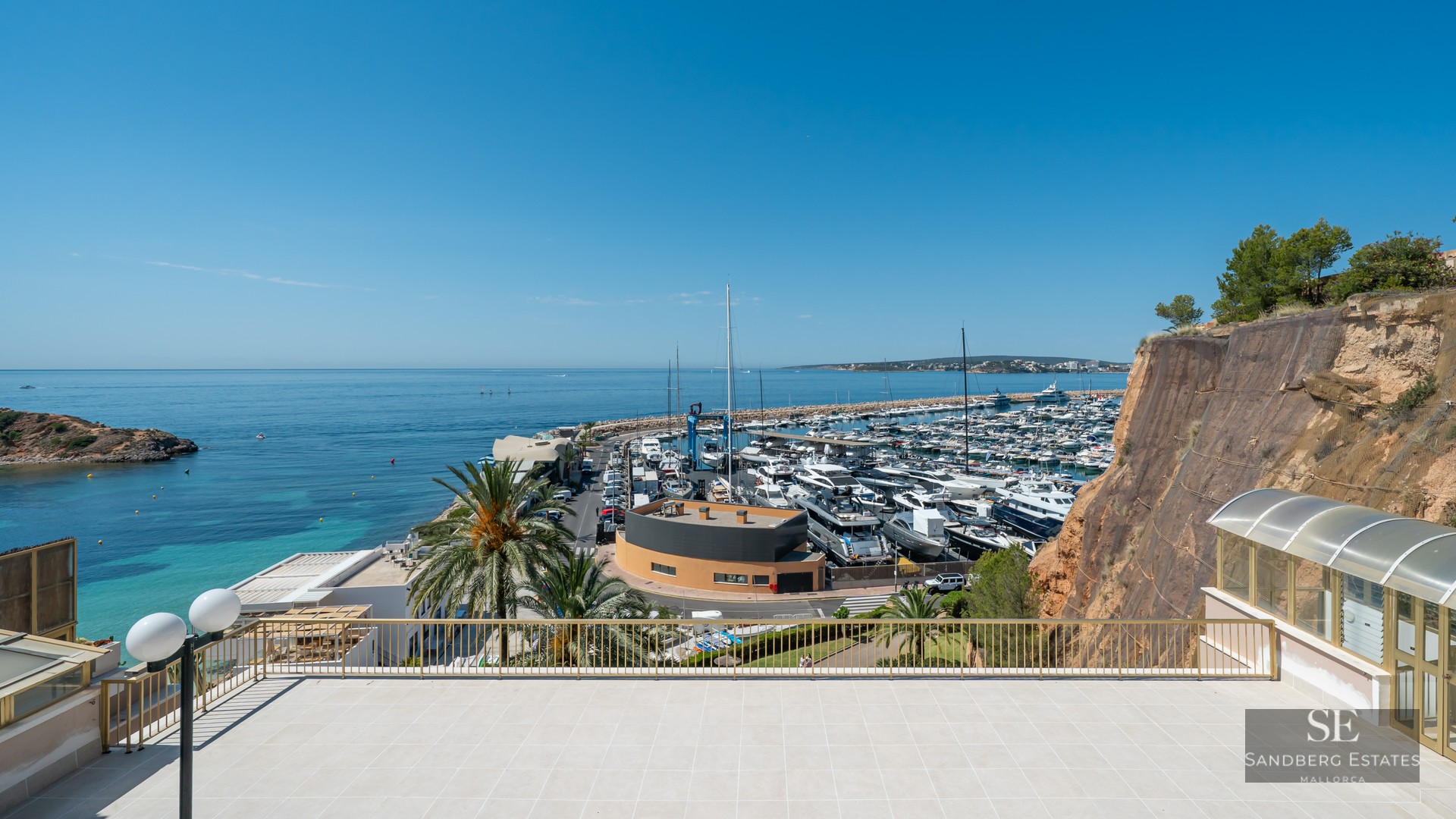 Elevated terrace with gold railing overlooking a busy yacht marina and the blue Mediterranean sea.