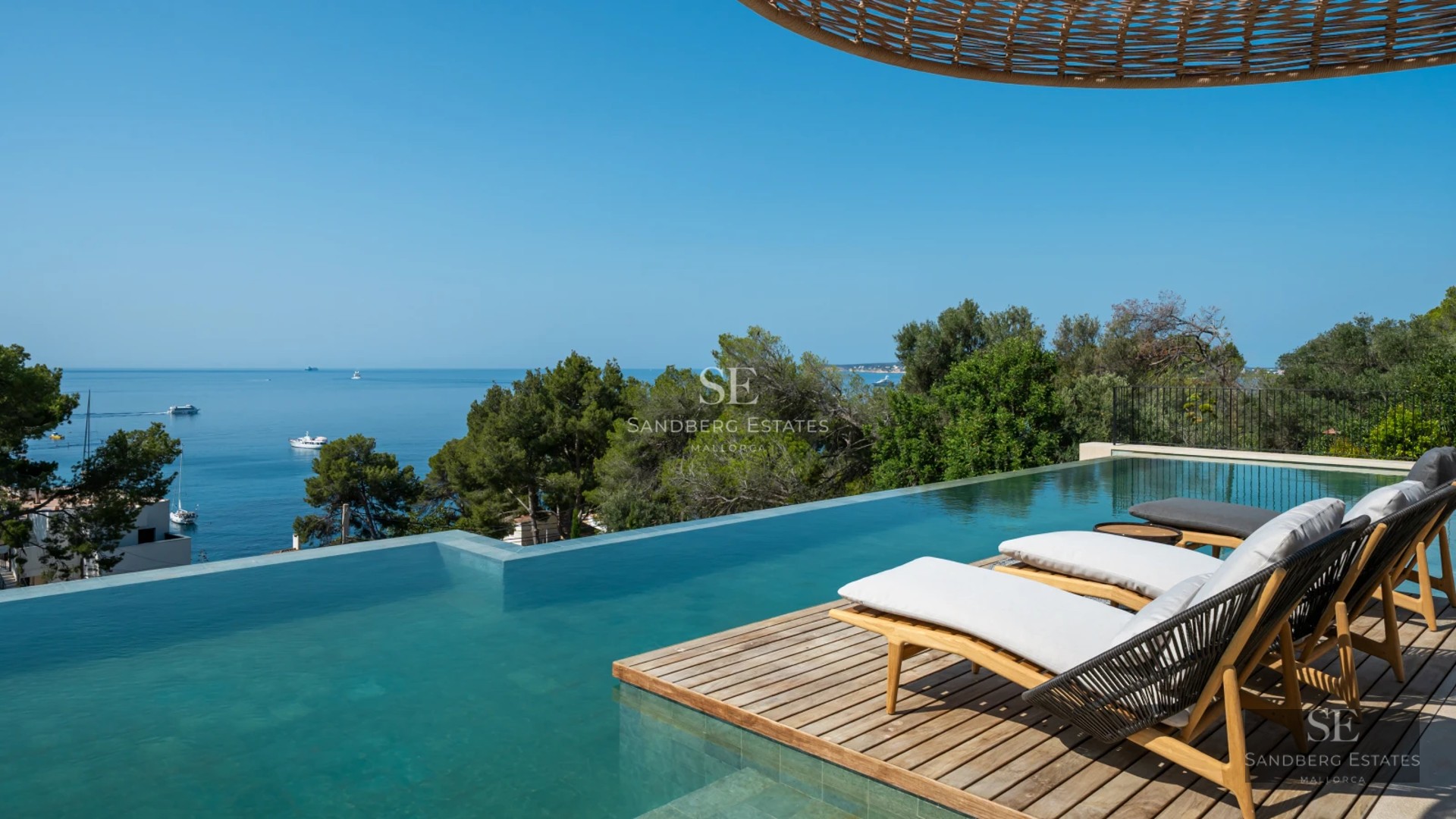 Infinity pool view with sun loungers, lush garden, and modern architecture. Large windows and blue sky.