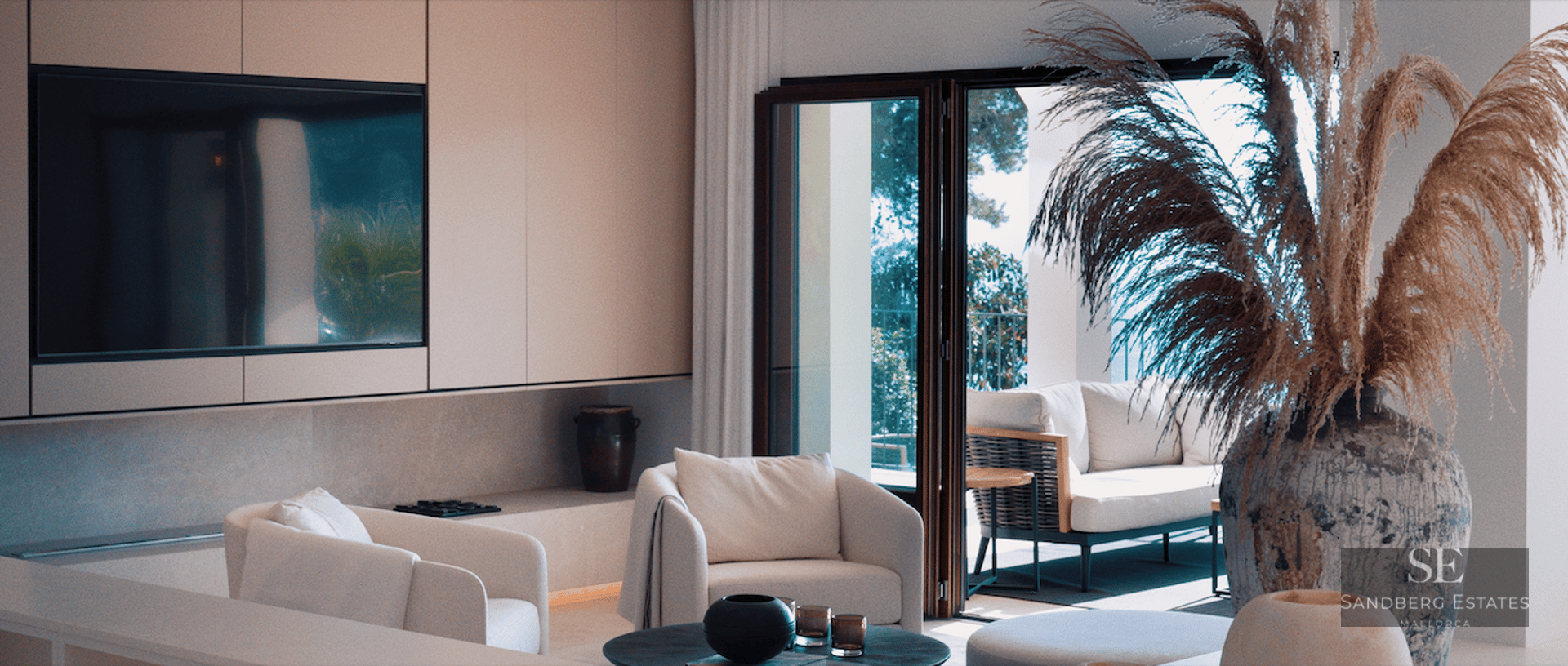 Bright minimalist living room with white armchairs, integrated TV, and large glass doors opening to a sunny terrace.