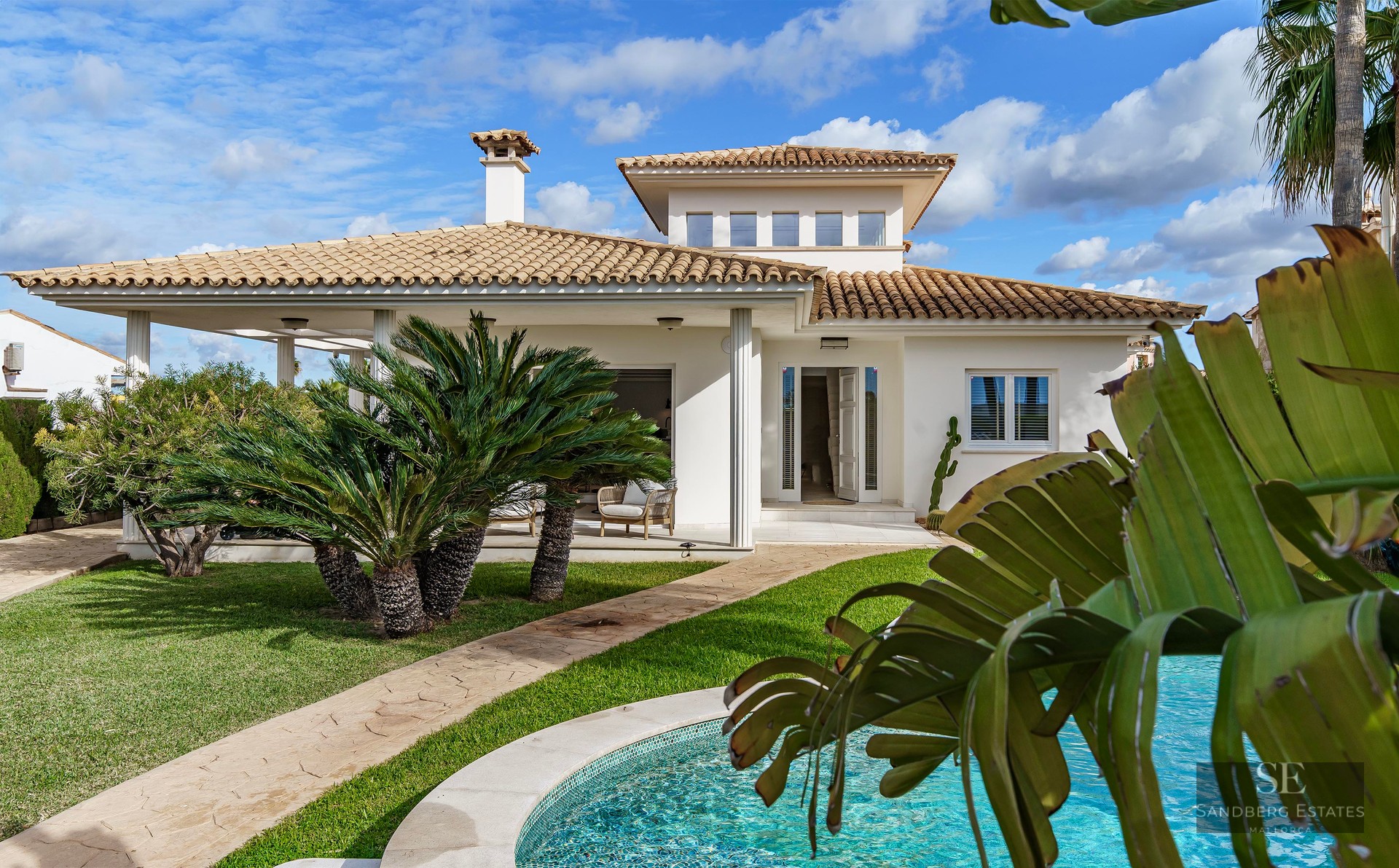 White Mediterranean villa with a terracotta roof, a lush green lawn, palm trees, and a turquoise swimming pool.