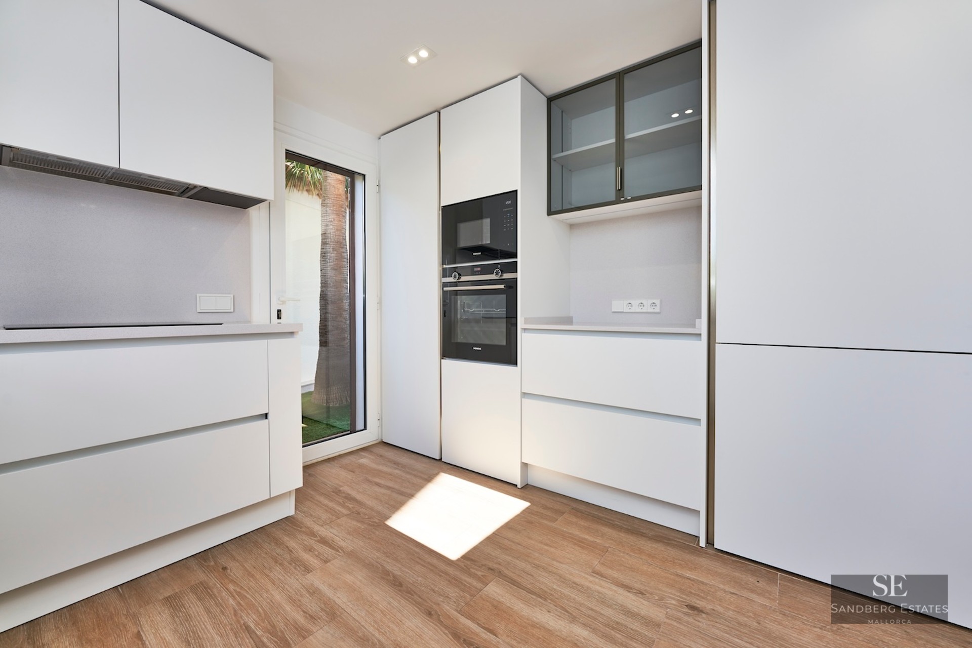 Minimalist white kitchen with integrated appliances, wood-look floors, and a glass door showing a palm tree.
