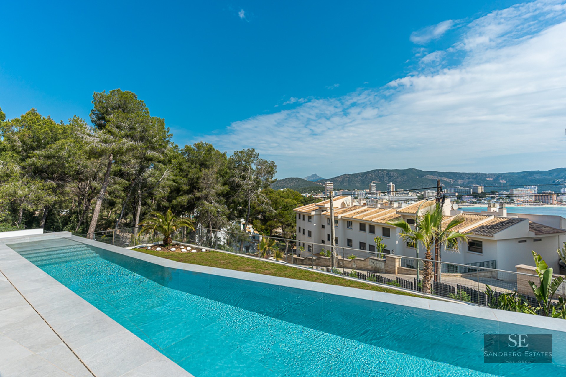 Luxury infinity swimming pool with stone tiling, overlooking a coastal town, mountains, and the sea under a clear blue sky.