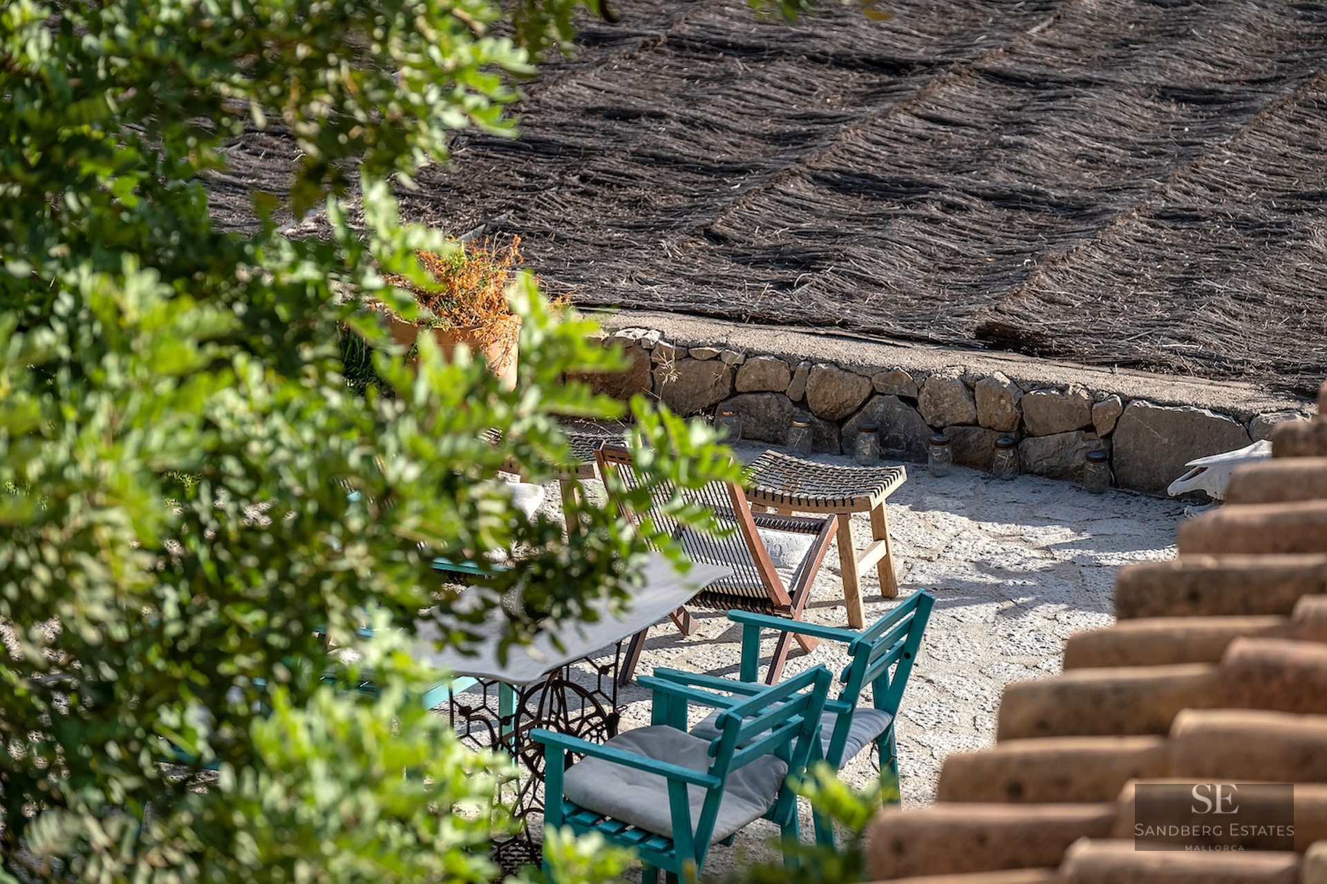 Outdoor stone terrace with teal wooden chairs, vintage table, and a thatched roof seen through greenery.