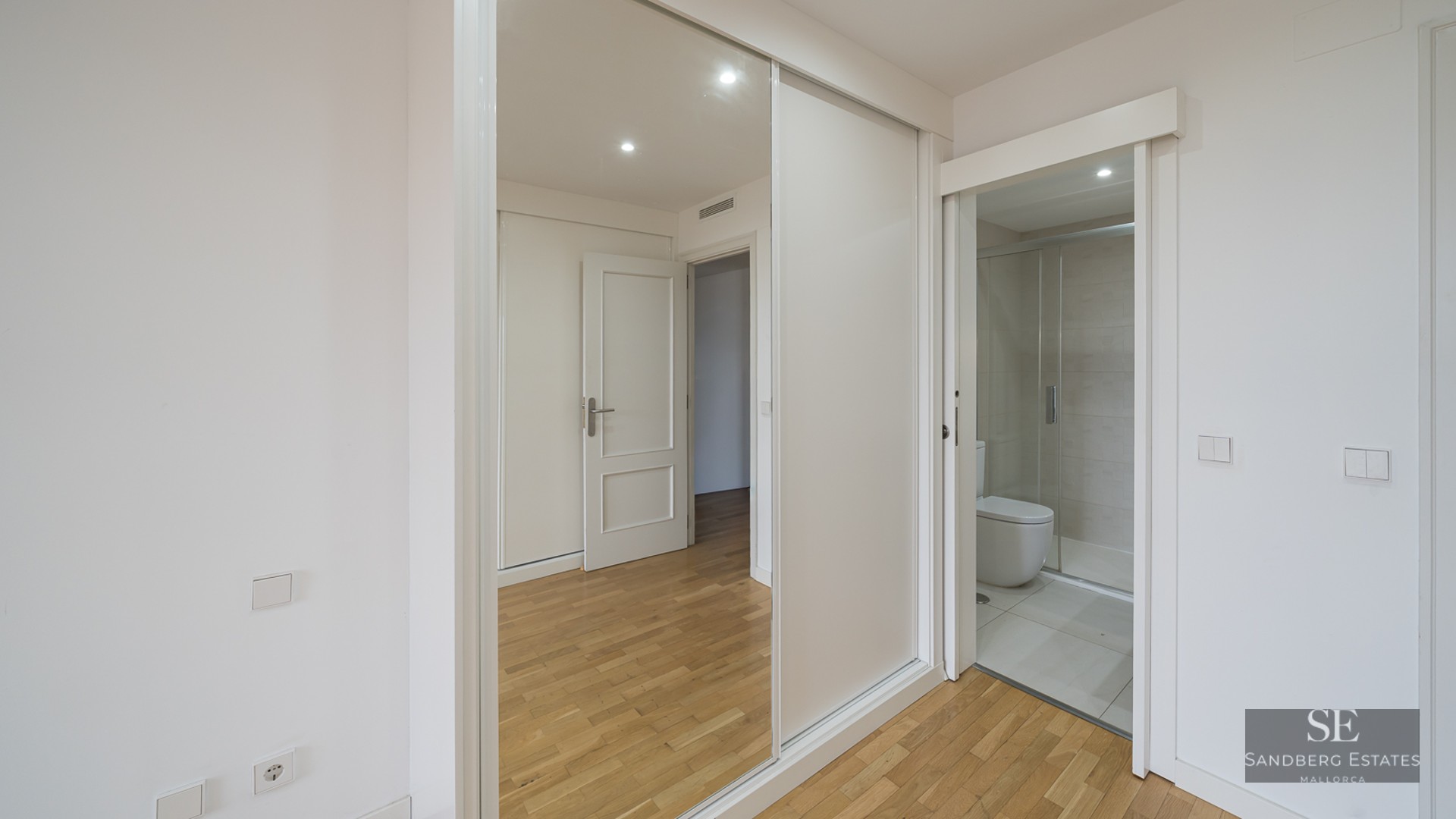 A bright modern room with light wood floors, a large mirrored sliding wardrobe, and a doorway leading into a white tiled bathroom.