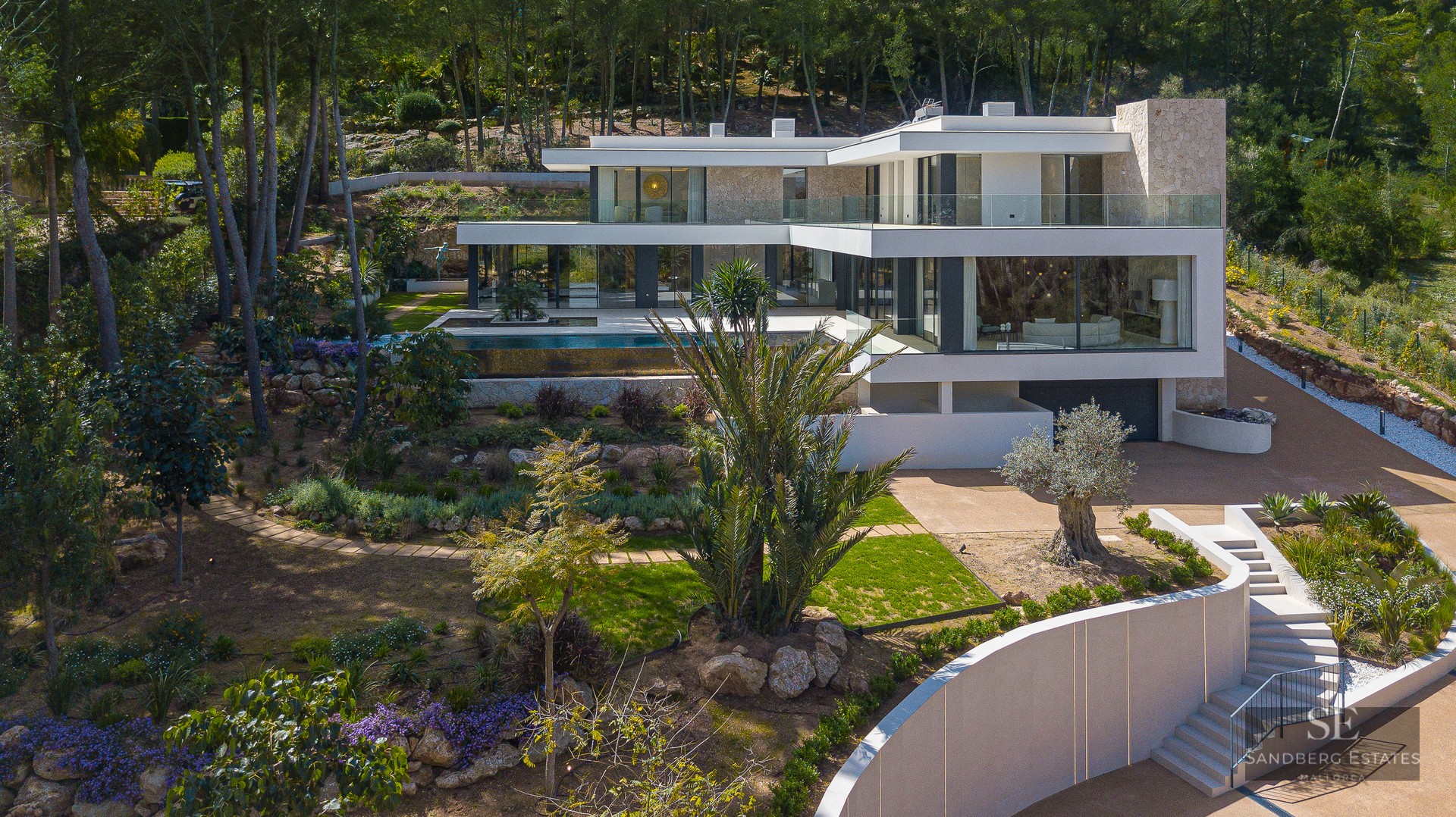 Aerial view of a three-story modern white villa with an infinity pool, landscaped gardens, and natural stone walls.