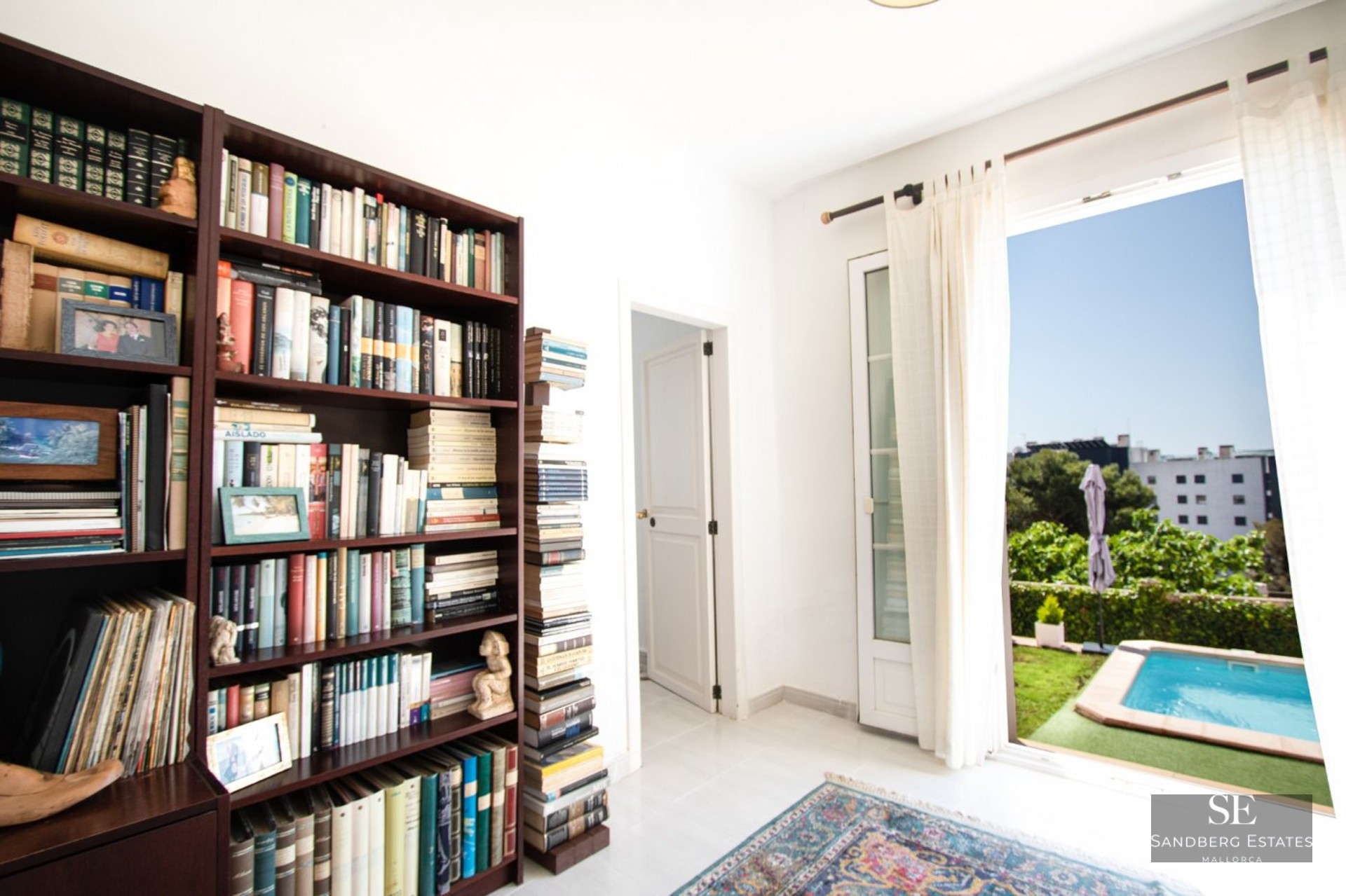A room featuring a large dark wood bookshelf filled with books and an open door leading to an outdoor swimming pool.