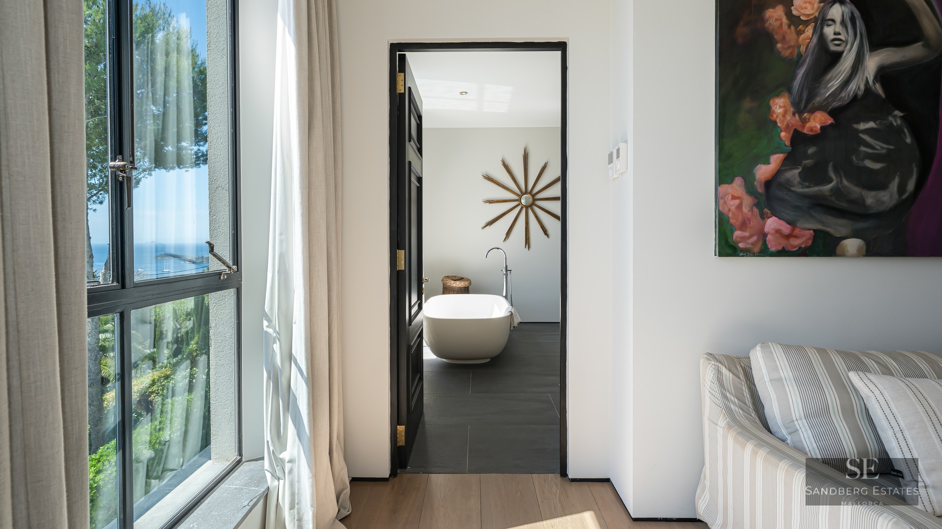 Open doorway revealing a modern bathroom with a freestanding tub and sunburst mirror, next to a window with an ocean view.