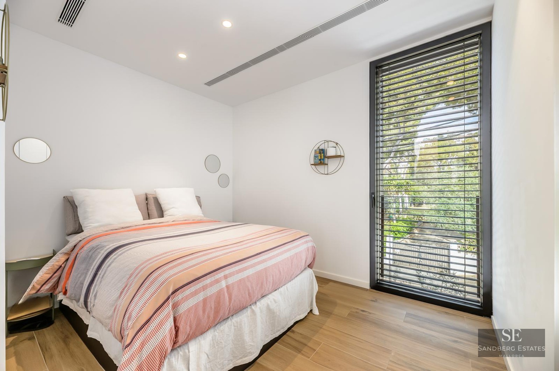 Bright bedroom featuring a striped bed, light wood flooring, and a large window looking out onto lush greenery.