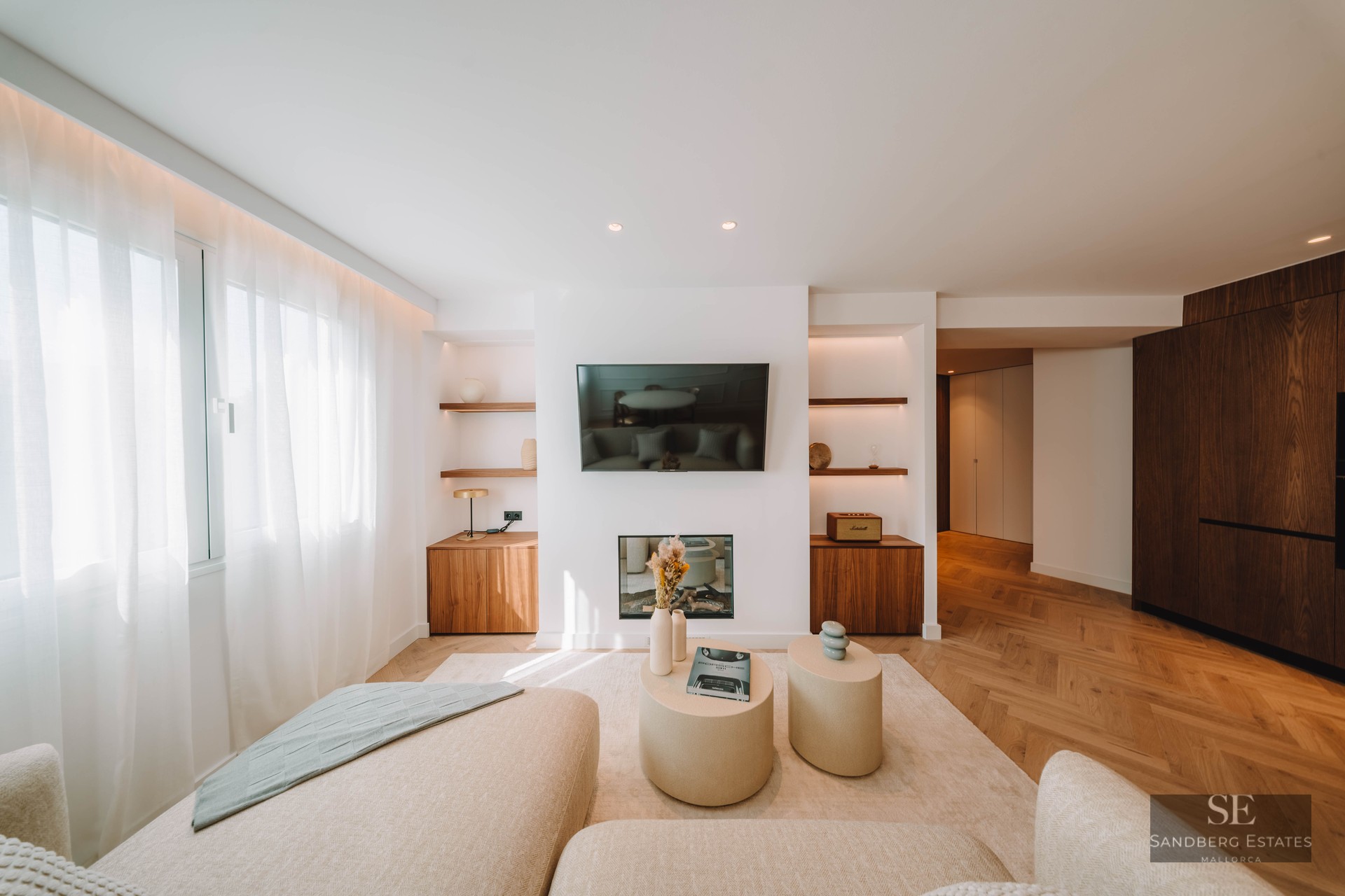 Bright living room featuring a wall-mounted TV, electric fireplace, herringbone wood floors, and cream sofas.