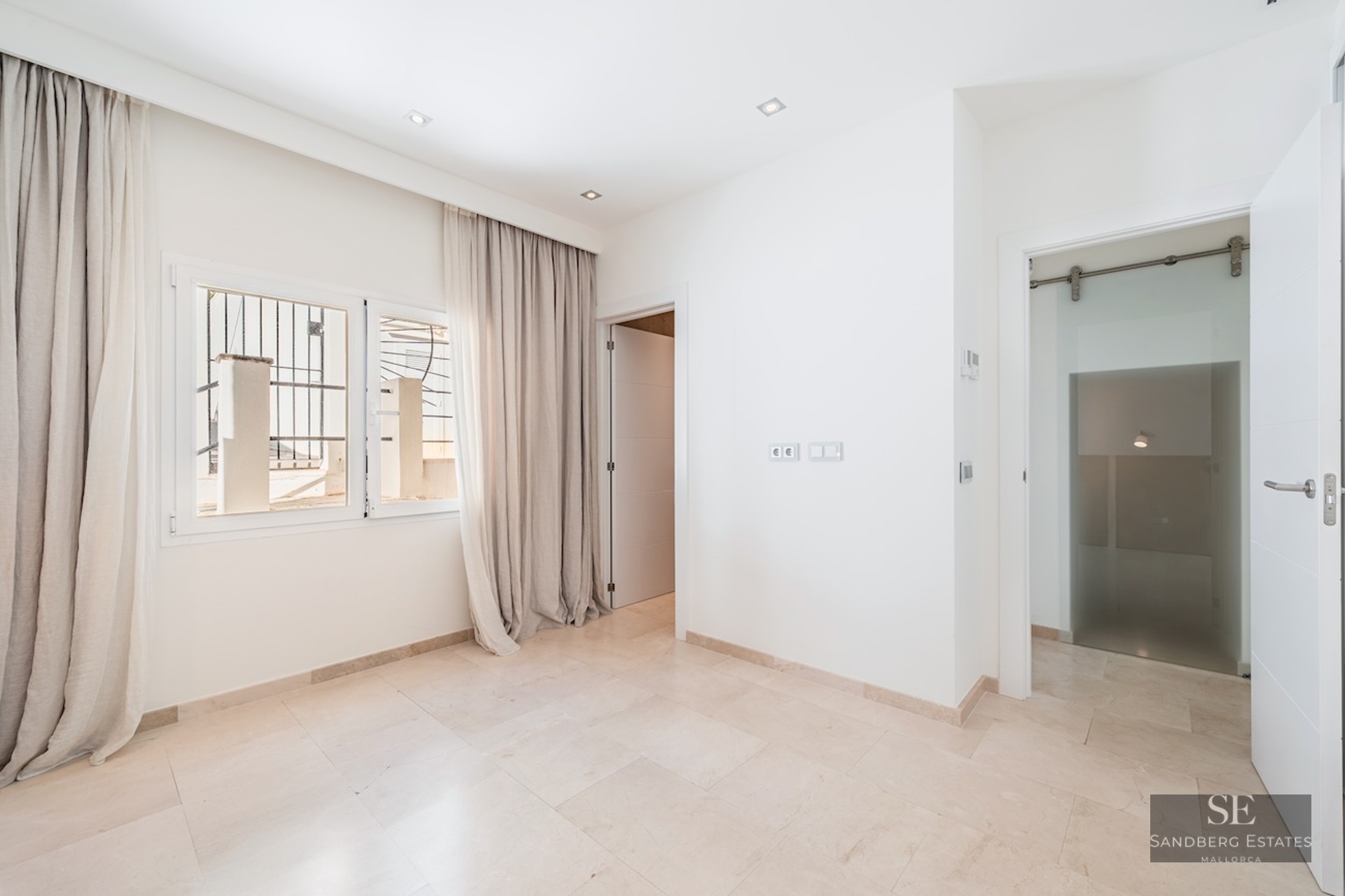 Bright empty room with white walls, cream marble floor, window with floor-length beige curtains, and open doorways.