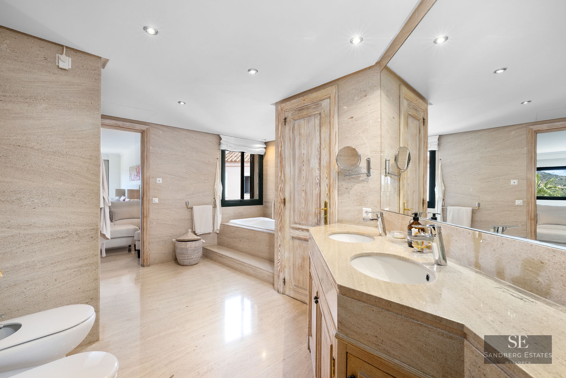 Salle de bain spacieuse avec murs en pierre beige, double vasque, grand miroir et baignoire encastrée au fond.