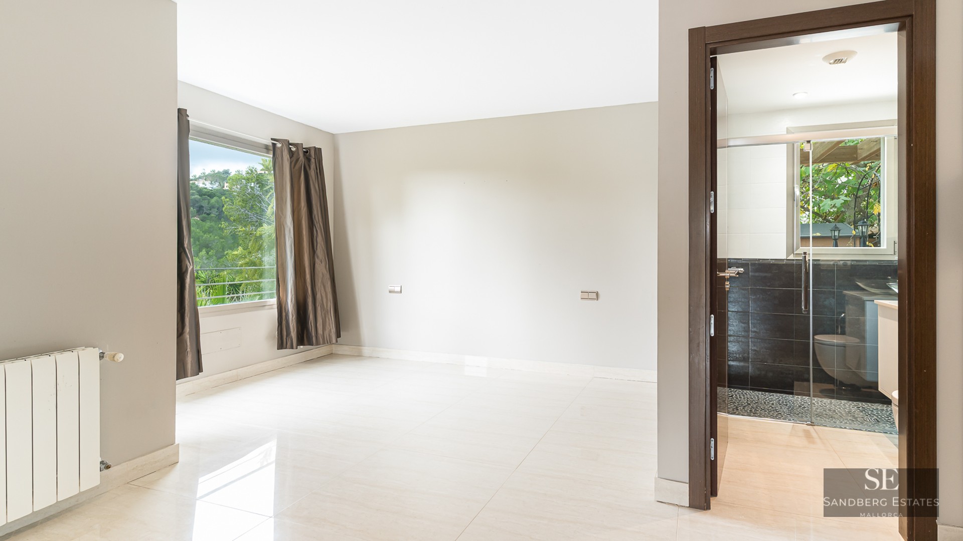 Empty modern bedroom with polished cream tile floors, a window with dark curtains, and an open doorway to an ensuite bathroom.