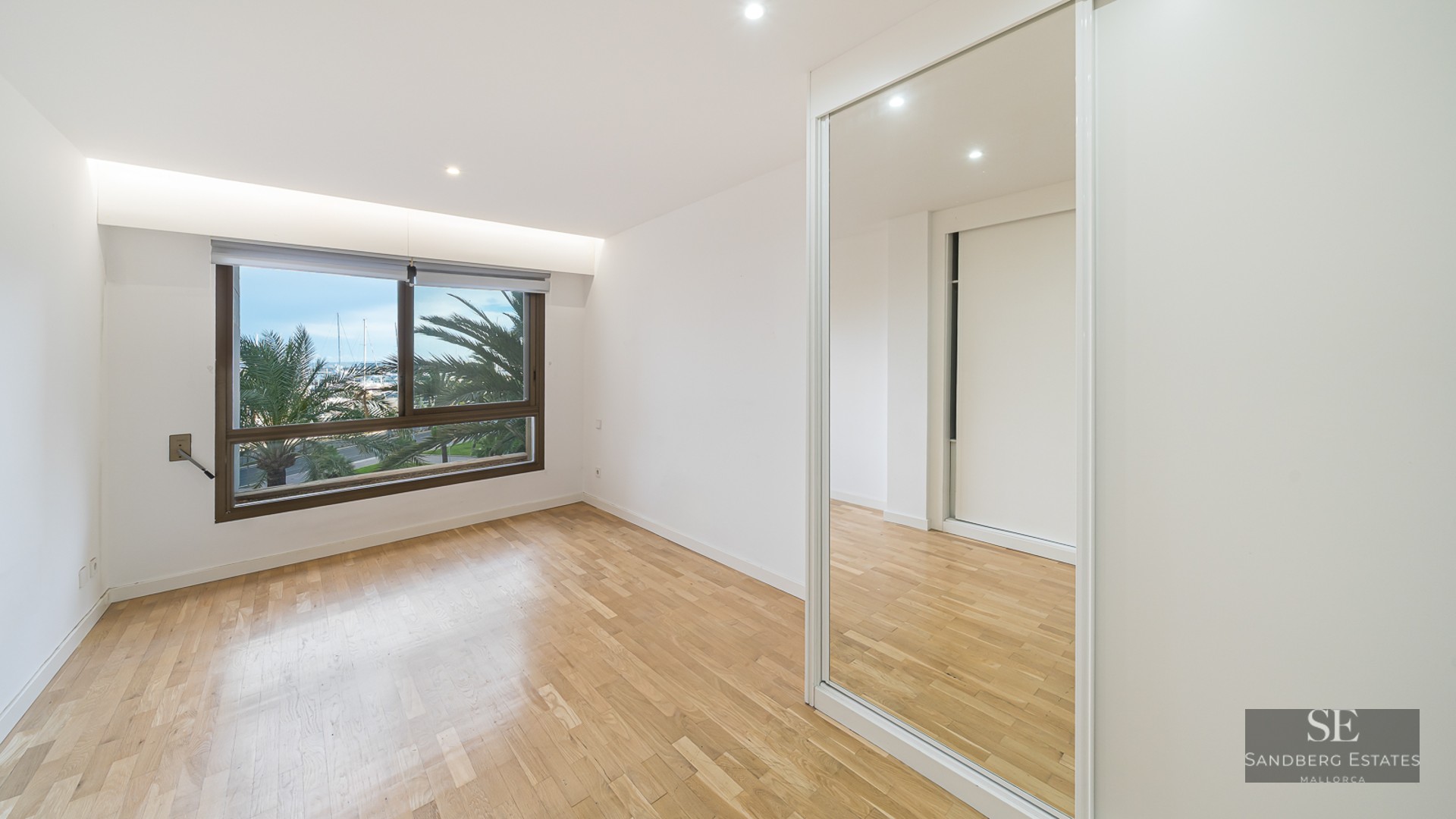 Empty bedroom with light wood floors, mirrored wardrobe, and a window showing palm trees and boats.