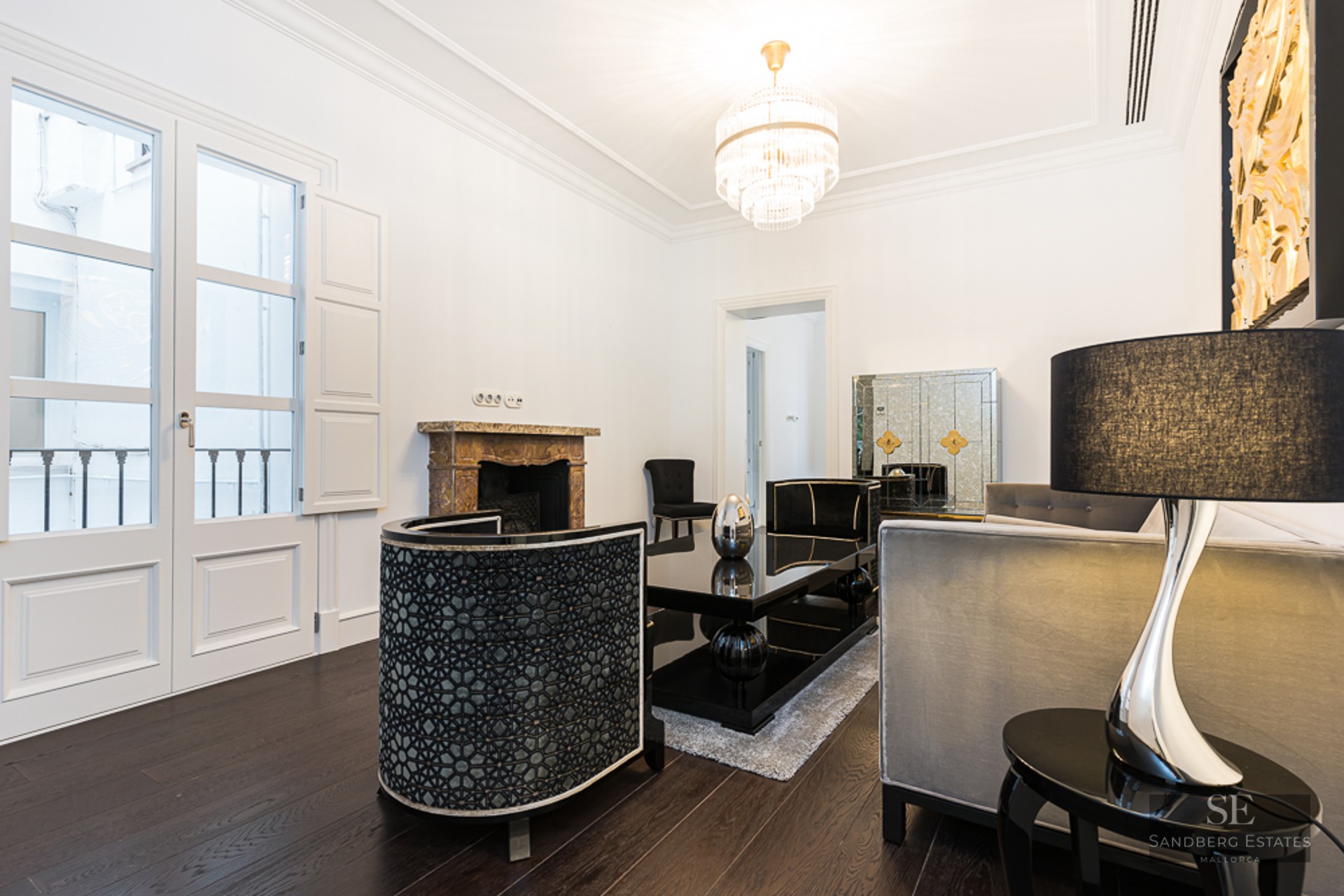Elegant living room with white walls, dark wood floors, designer furniture, crystal chandelier, and a marble fireplace.