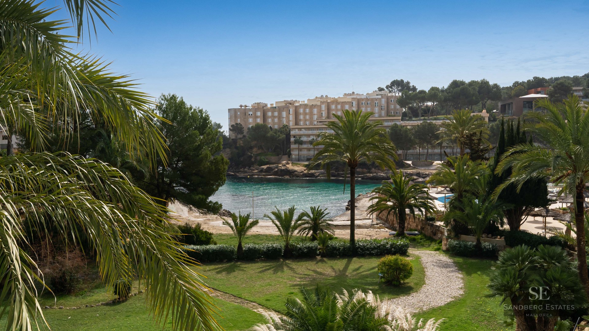 Lush garden with stone path and palm trees overlooking a turquoise sea cove and coastal buildings under a clear blue sky.