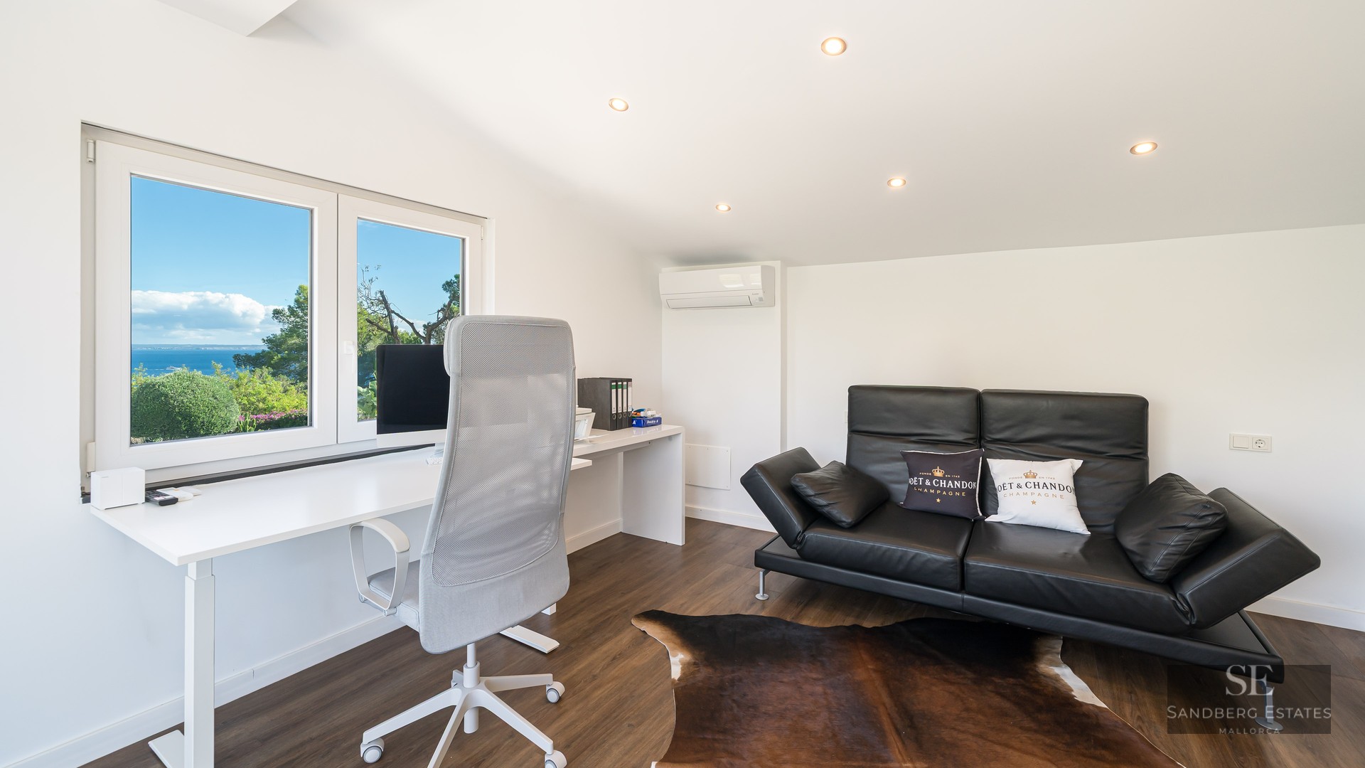 Bureau lumineux avec table blanche, chaise ergonomique et canapé en cuir noir devant une fenêtre donnant sur la mer.
