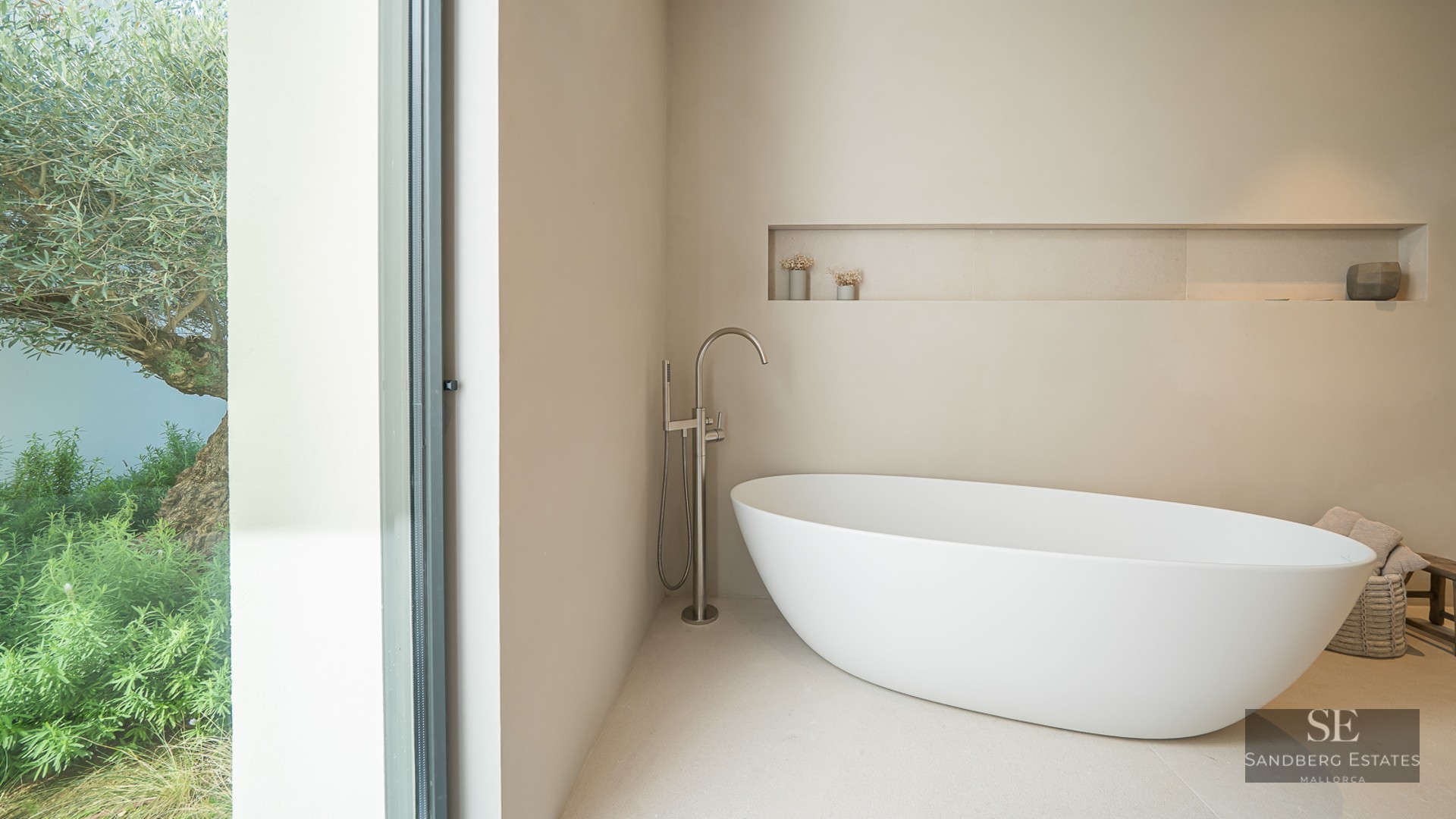 Minimalist bathroom featuring a white freestanding bathtub next to a floor-to-ceiling window with a garden view.