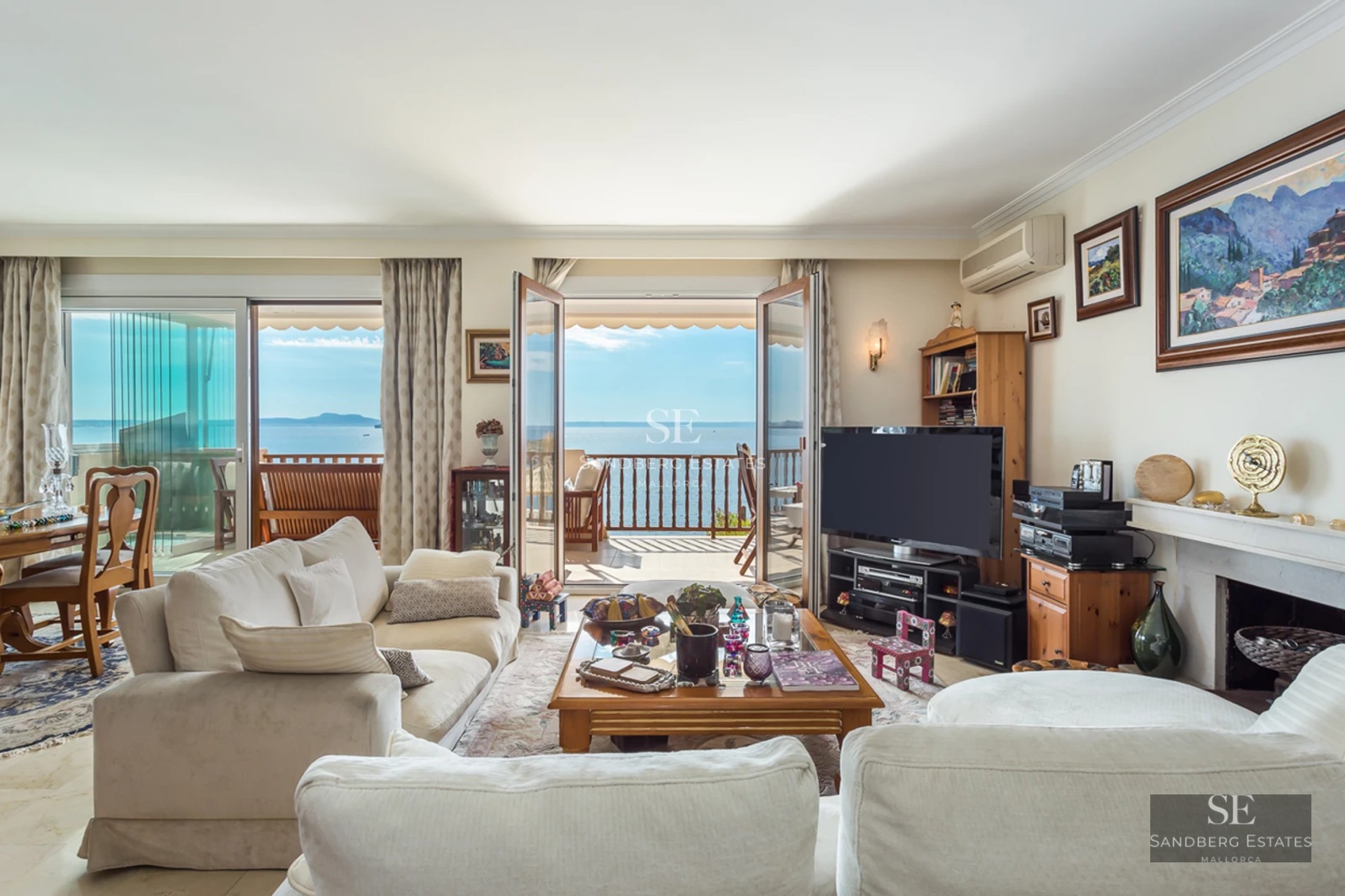 Bright living room with white sofas, wooden furniture, and glass doors opening to a balcony with an ocean view.