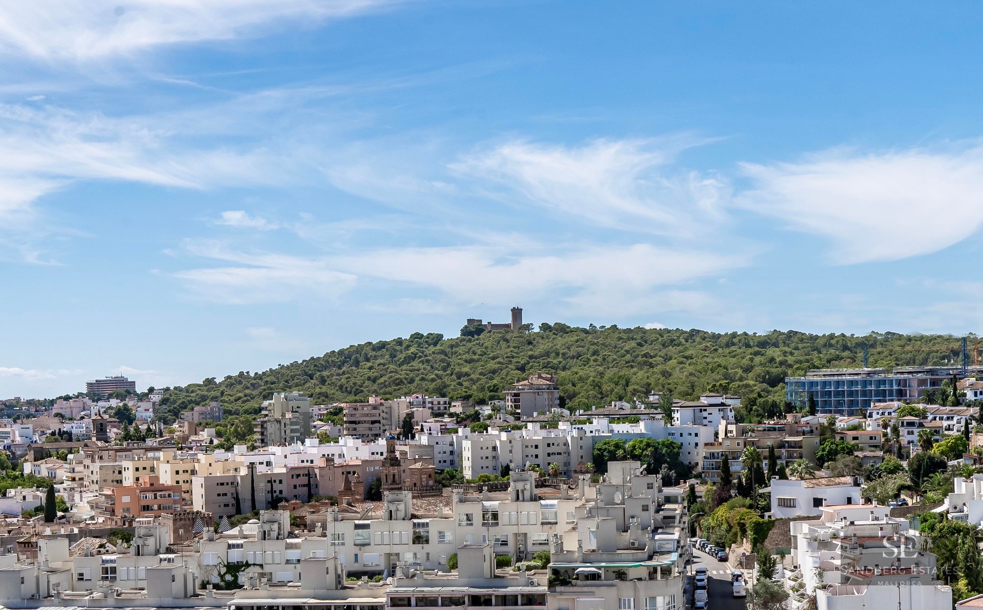 Panoramic view of Palma featuring white buildings and the historic Bellver Castle on a lush green hill.