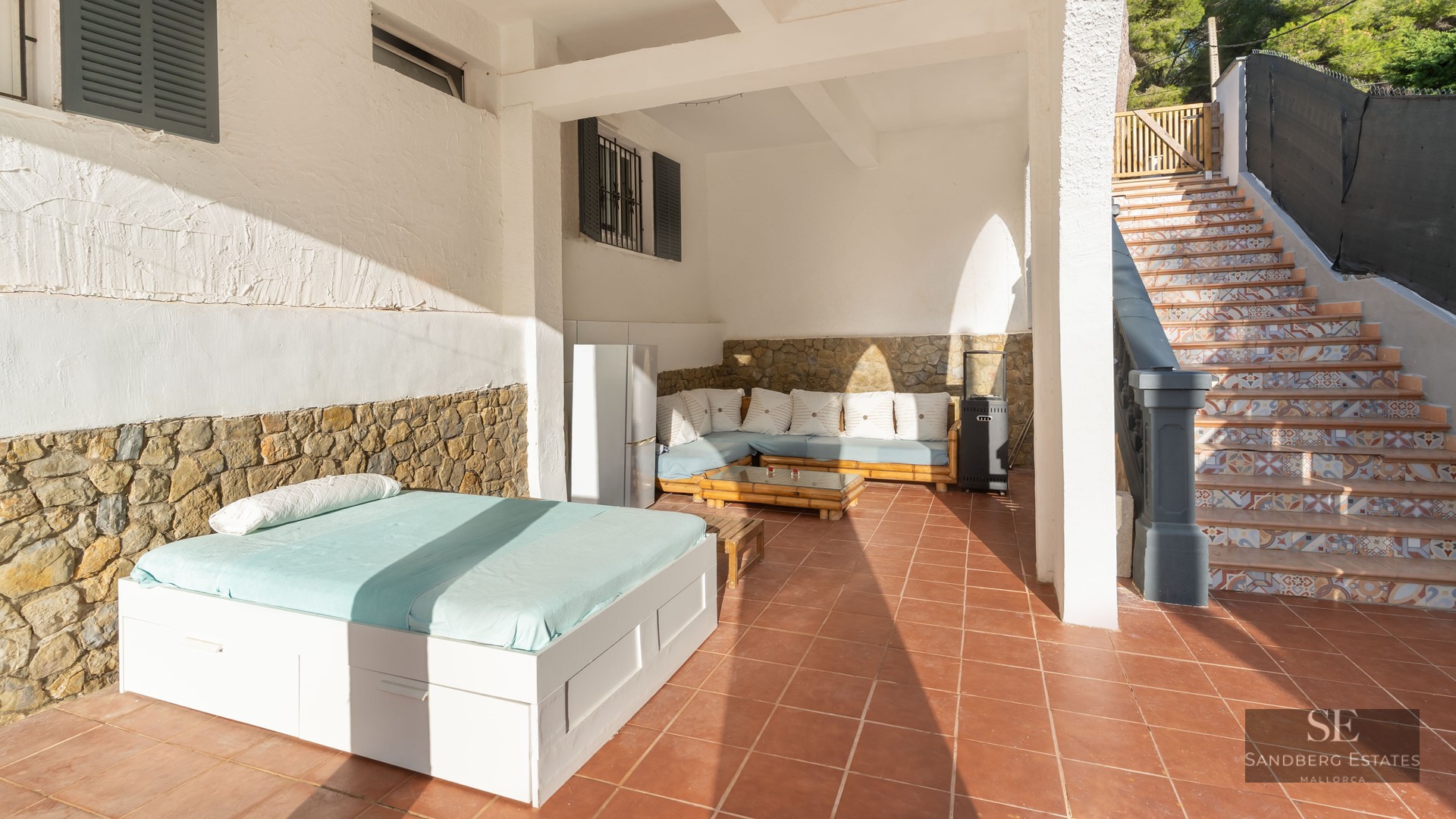 Terrasse couverte avec sols en terre cuite, lit de jour extérieur, banquette, murs en pierre et escalier carrelé.