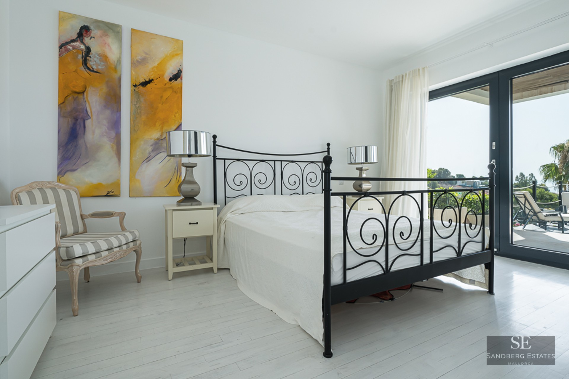 Bright white bedroom featuring a black wrought iron bed, abstract art, and large glass doors to a sunny terrace.