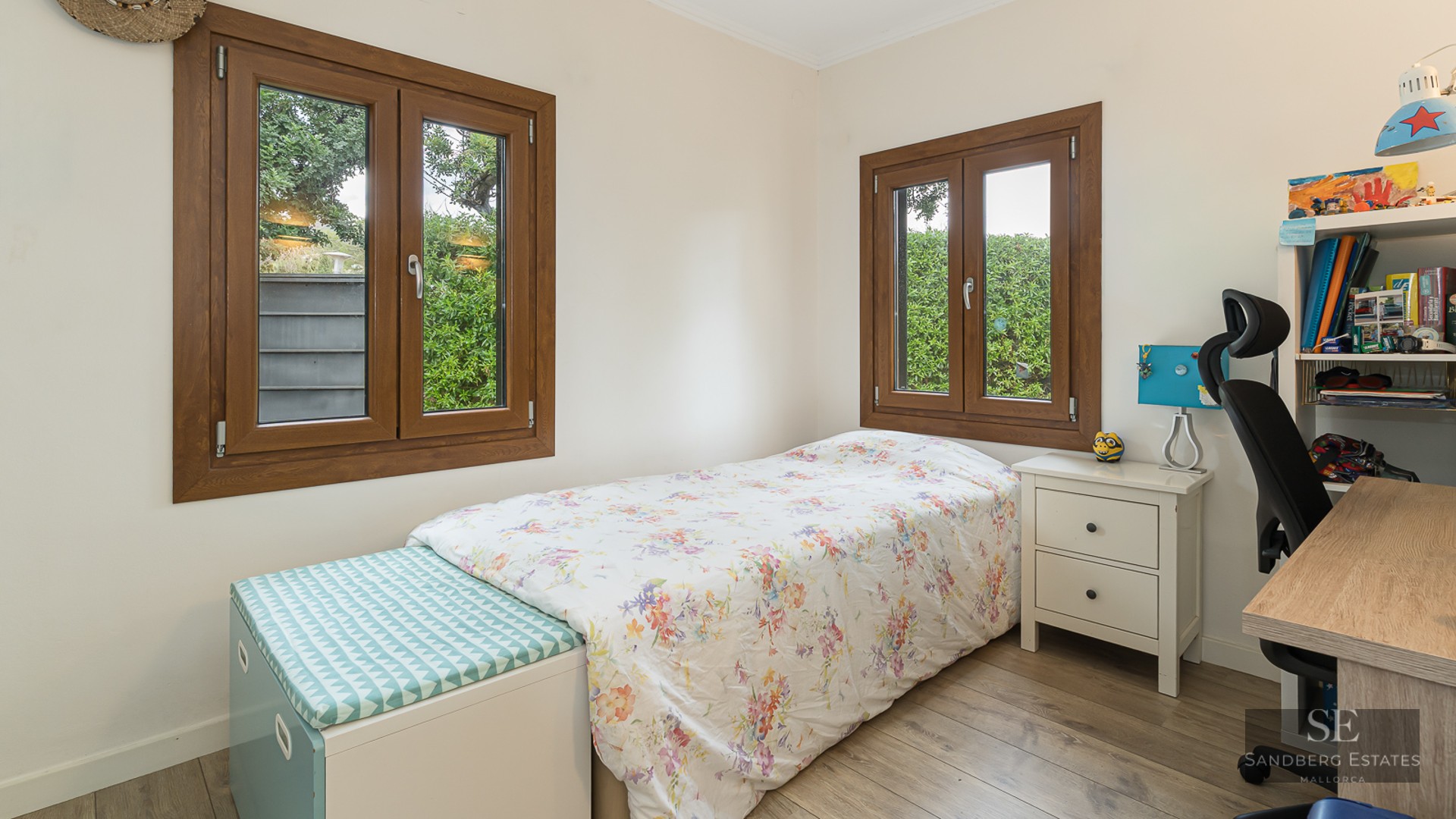 Bright bedroom with a single bed, desk, office chair, and wooden windows overlooking a green garden.