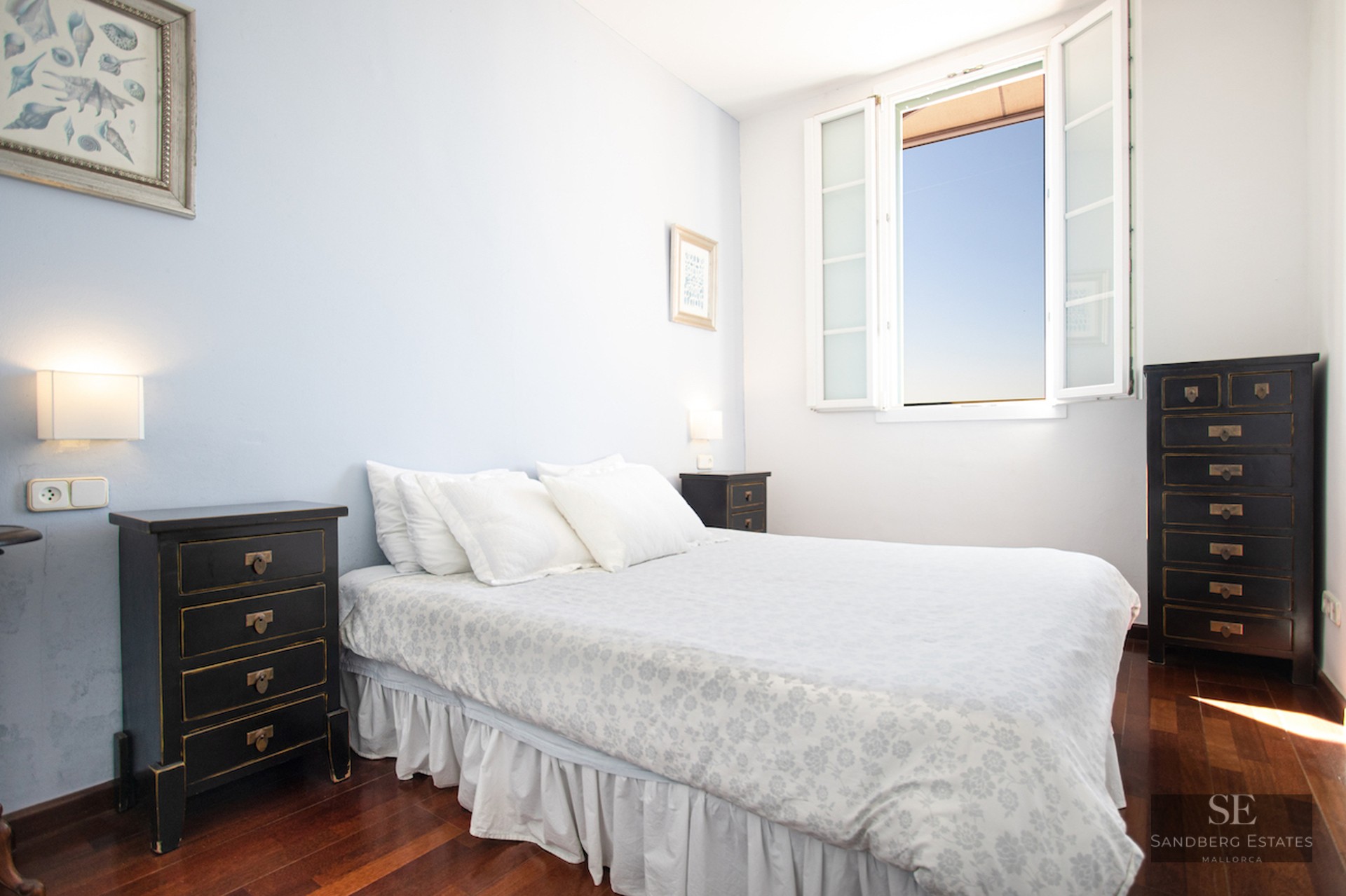 A bedroom with a white floral bed, black nightstands, dark wood floors, and an open window showing a blue sky.