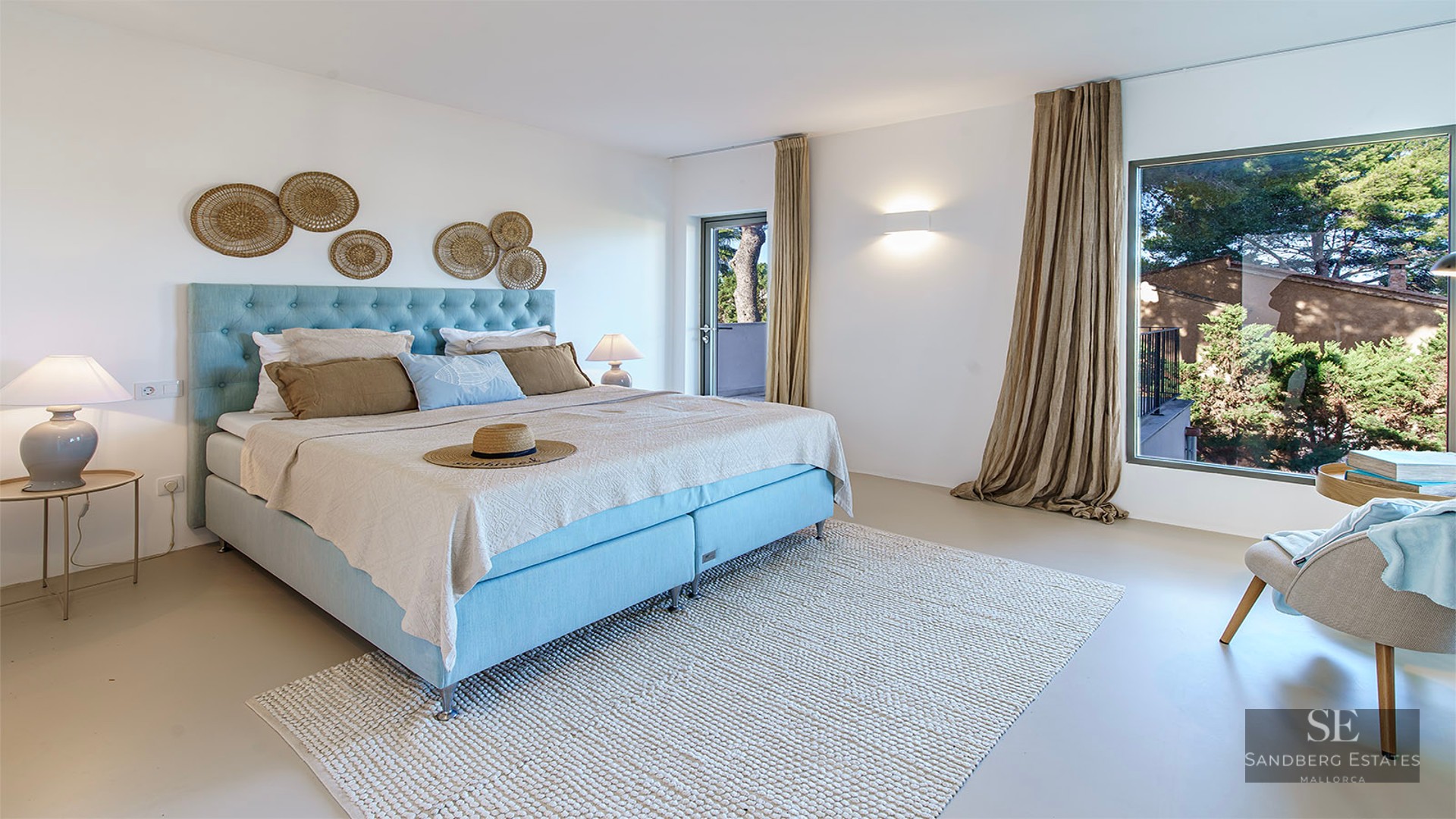 Bright room with light blue bed, wool rug, and large window overlooking green trees.