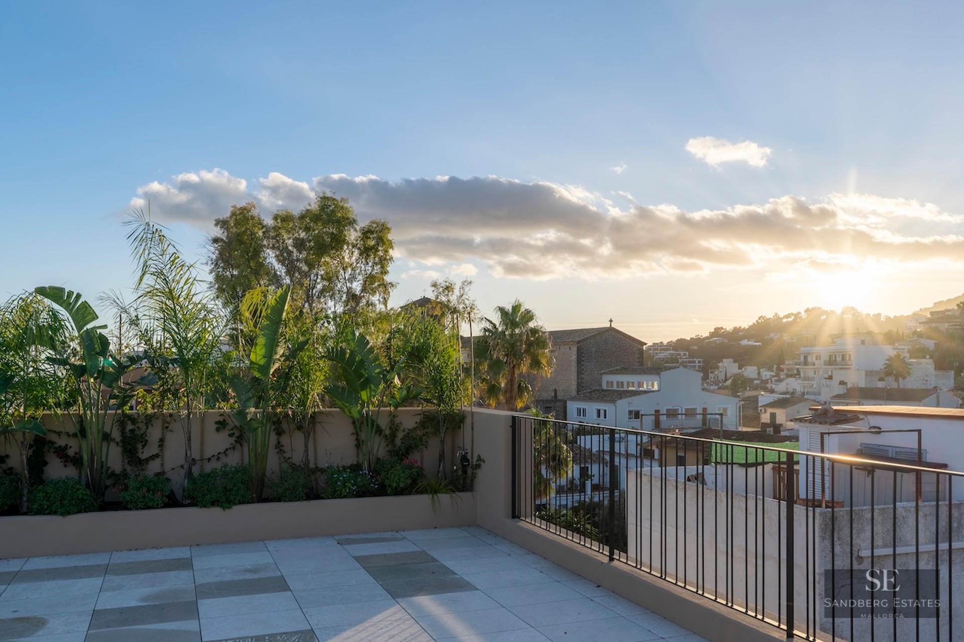Terrasse de toit moderne avec carrelage en pierre et plantes tropicales, surplombant une ville méditerranéenne au coucher du soleil.