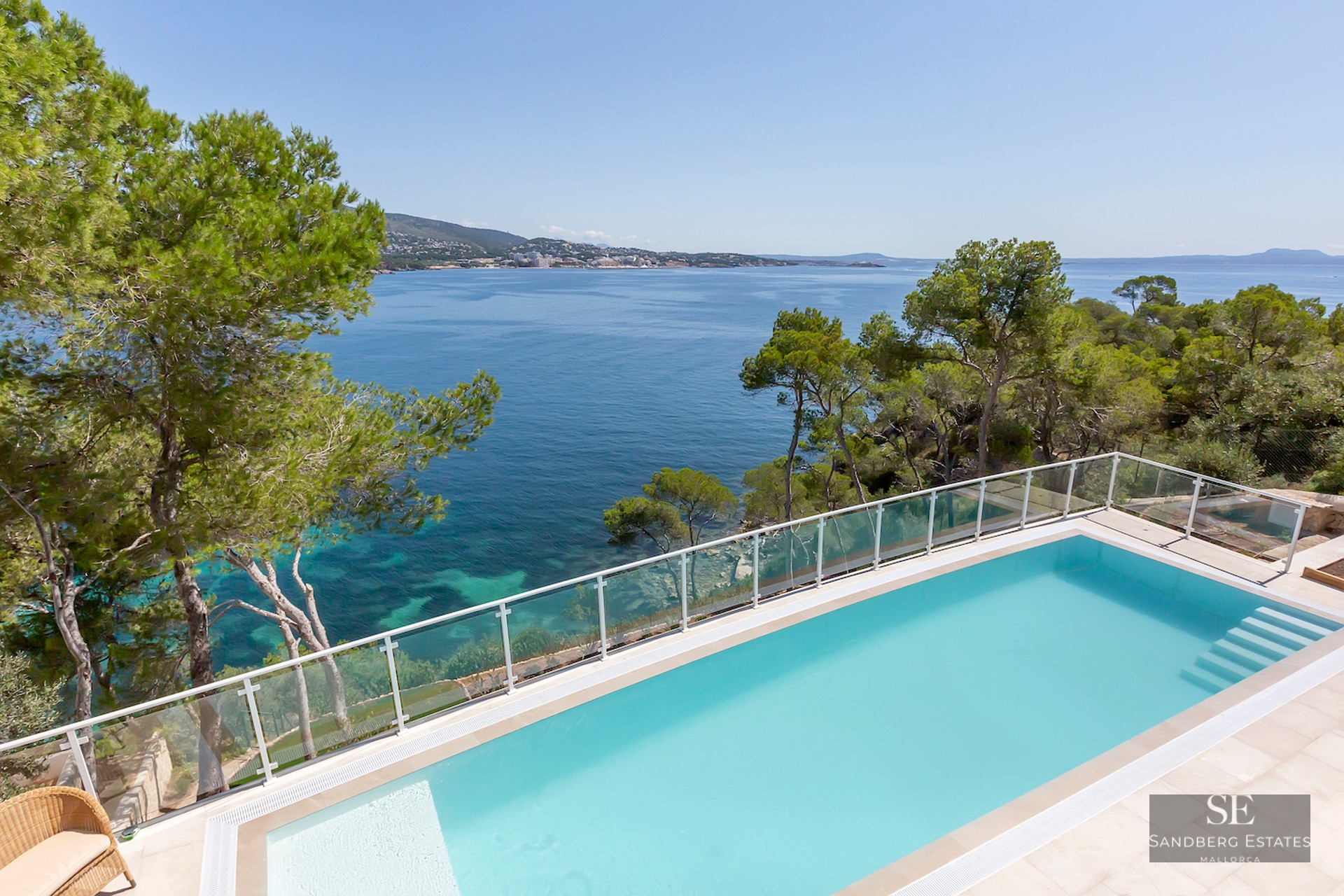 Rectangular turquoise swimming pool overlooking the Mediterranean sea with pine trees and a glass railing.