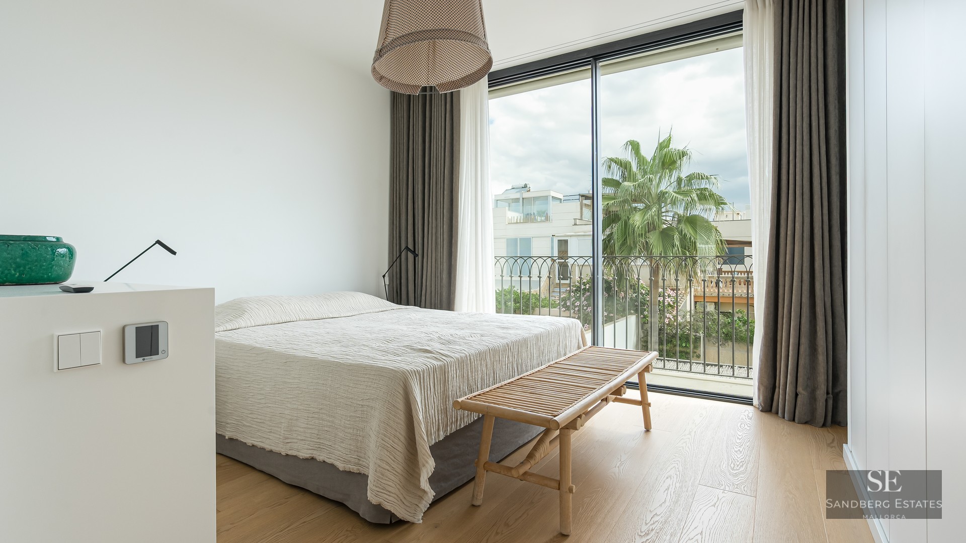 Bright bedroom with floor-to-ceiling windows, light wood floors, a large bed, and a palm tree view outside.