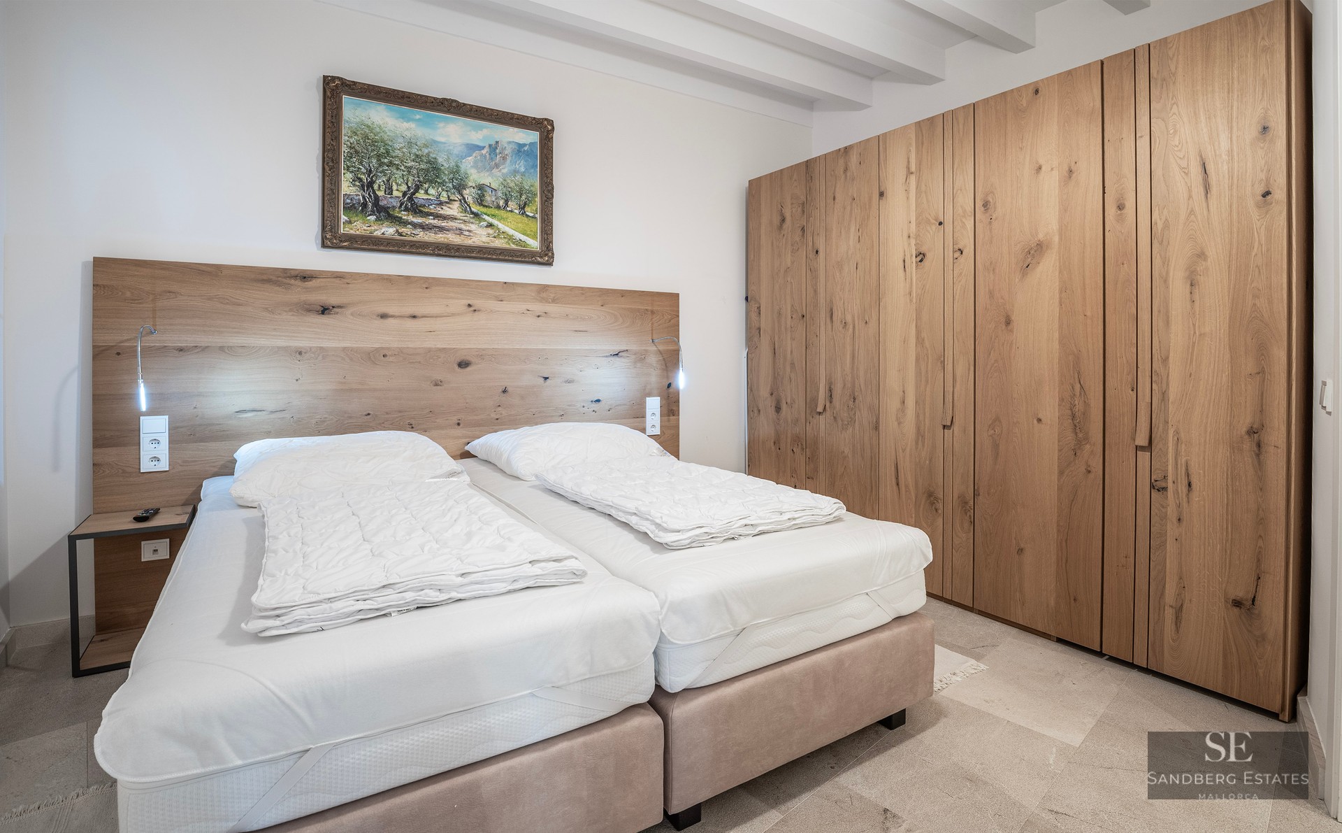 Bright bedroom with white beamed ceiling, large oak closet, and two beds with a wooden headboard.