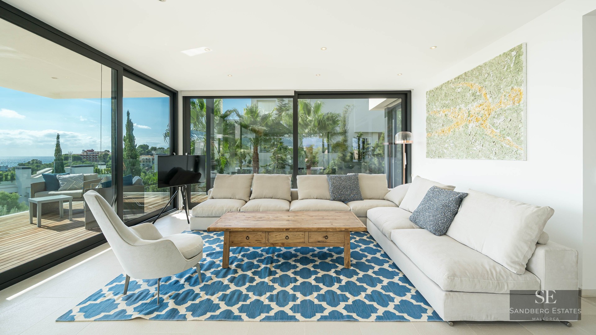 Modern living room with white L-shaped sofa, blue patterned rug, and floor-to-ceiling windows showing a coastal view.