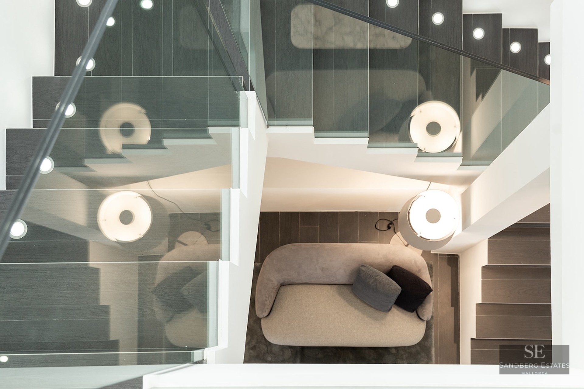 Overhead view of a staircase with glass railings looking down onto a grey sofa and a designer lamp.