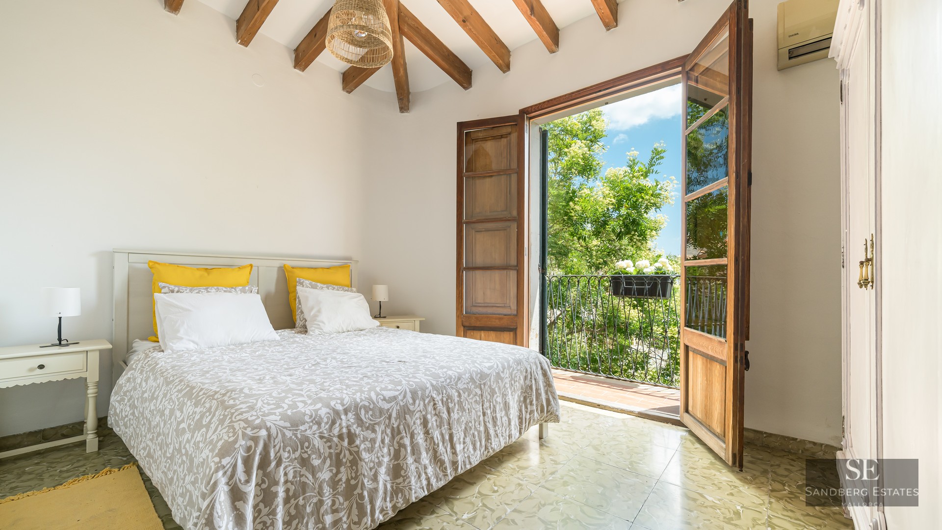 A bright bedroom with exposed wooden ceiling beams and French doors opening to a sunny balcony with green trees.