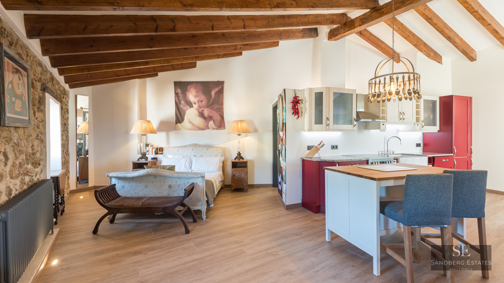 Studio featuring a classic bed, exposed wooden ceiling beams, a stone wall, and a modern red and white kitchen with island.