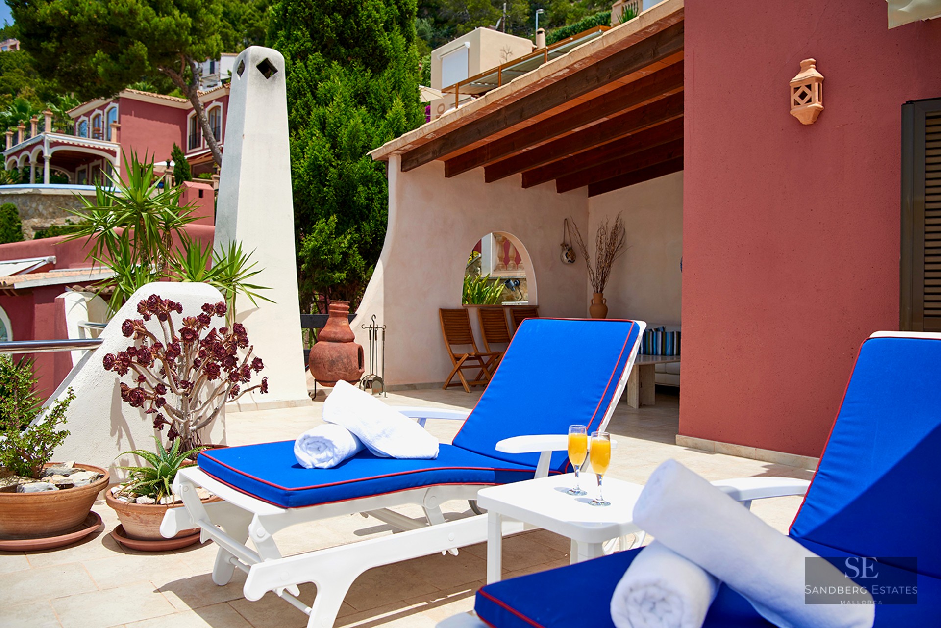 Sunny terrace featuring vibrant blue sun loungers, white towels, and a wooden-beamed porch.