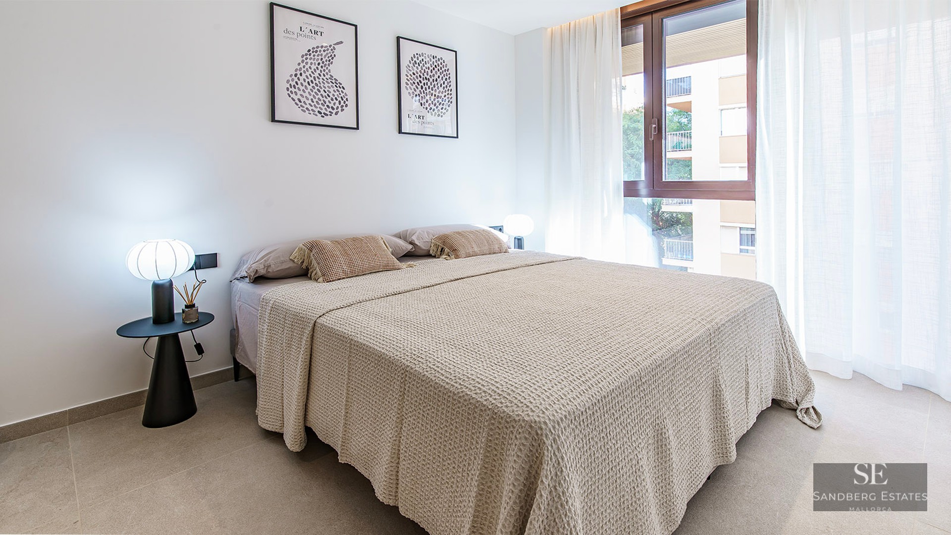 Bright bedroom with a large bed, beige waffle-knit linens, designer lamps, and a window with sheer white curtains.