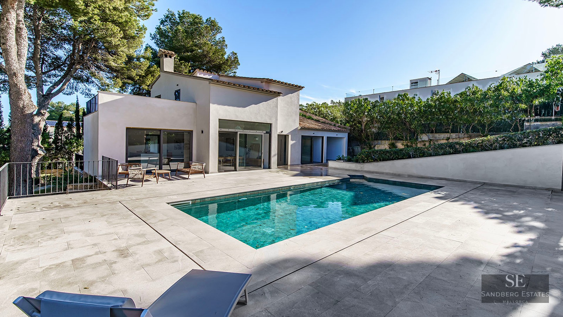 Modern white villa with a turquoise rectangular swimming pool and stone terrace surrounded by pine trees.