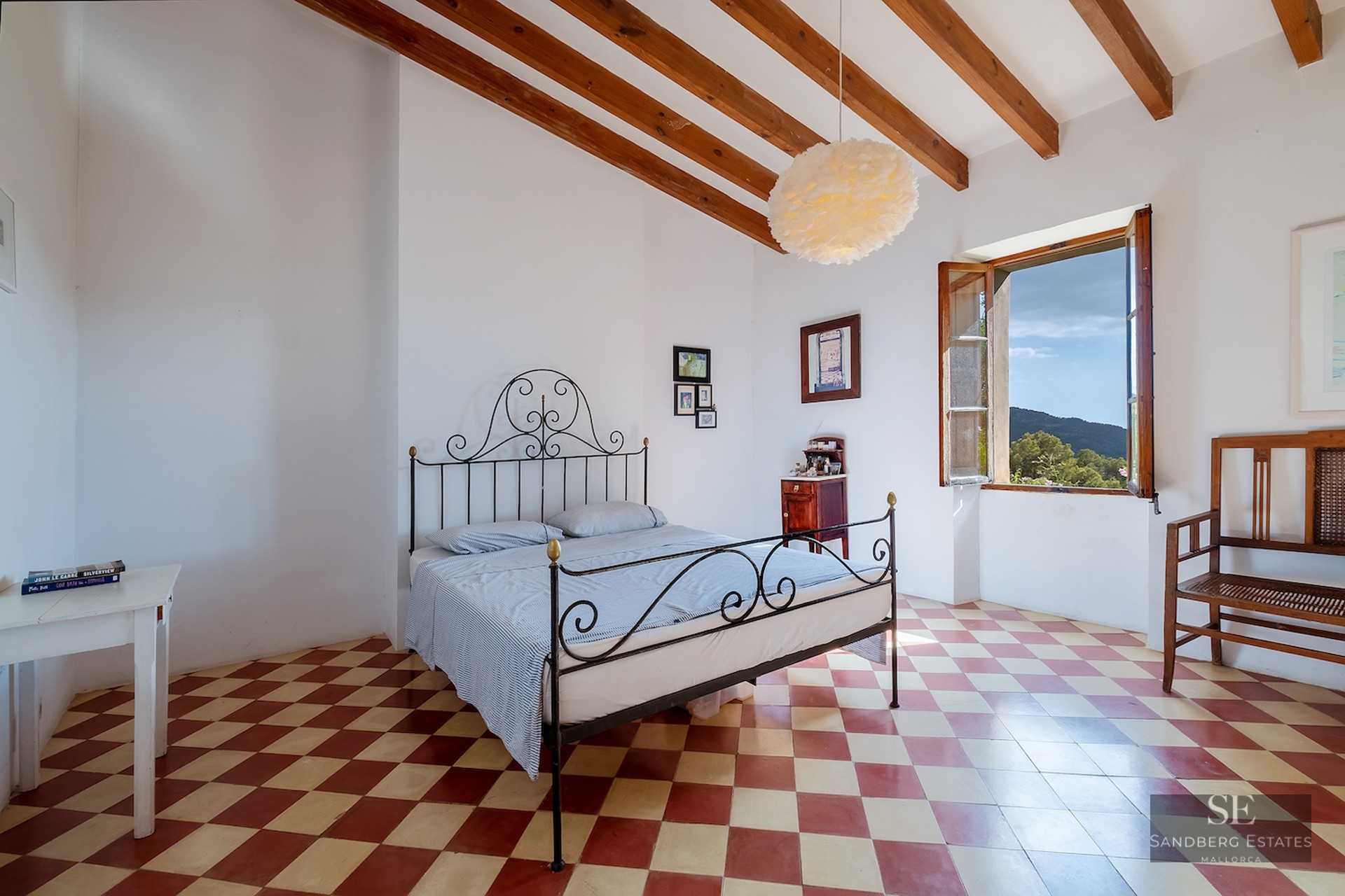 Bedroom with a black wrought iron bed, red and white checkered floor, exposed wooden beams, and a window with hill views.