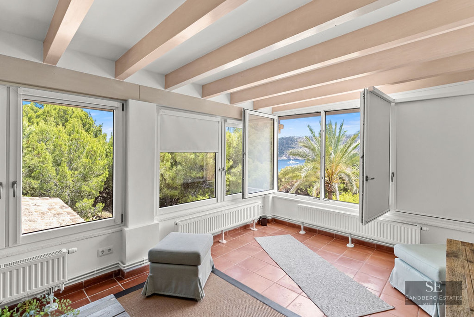 Interior room with terracotta floor tiles, wooden ceiling beams, and large windows overlooking pine trees and the sea.