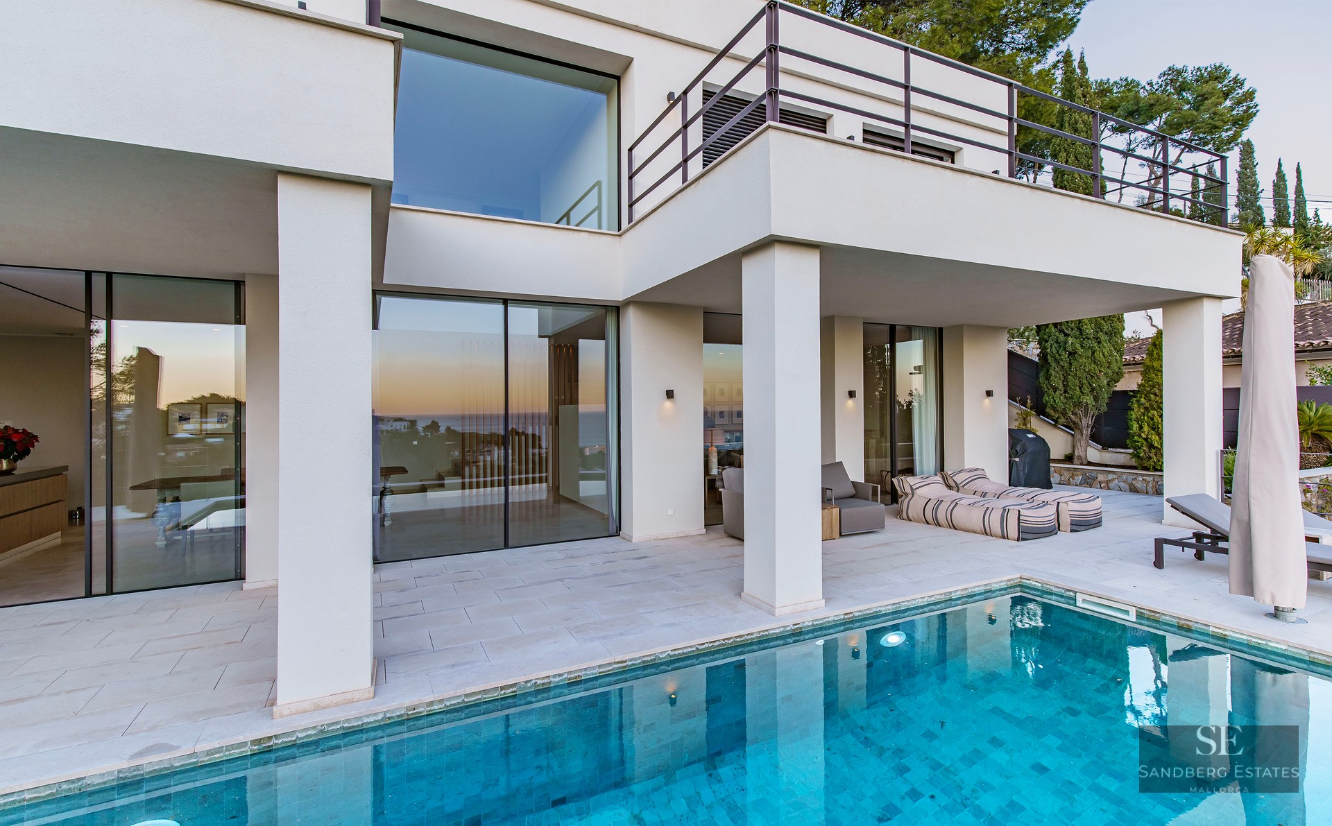 Modern white villa with a turquoise swimming pool and large glass windows reflecting a sunset.