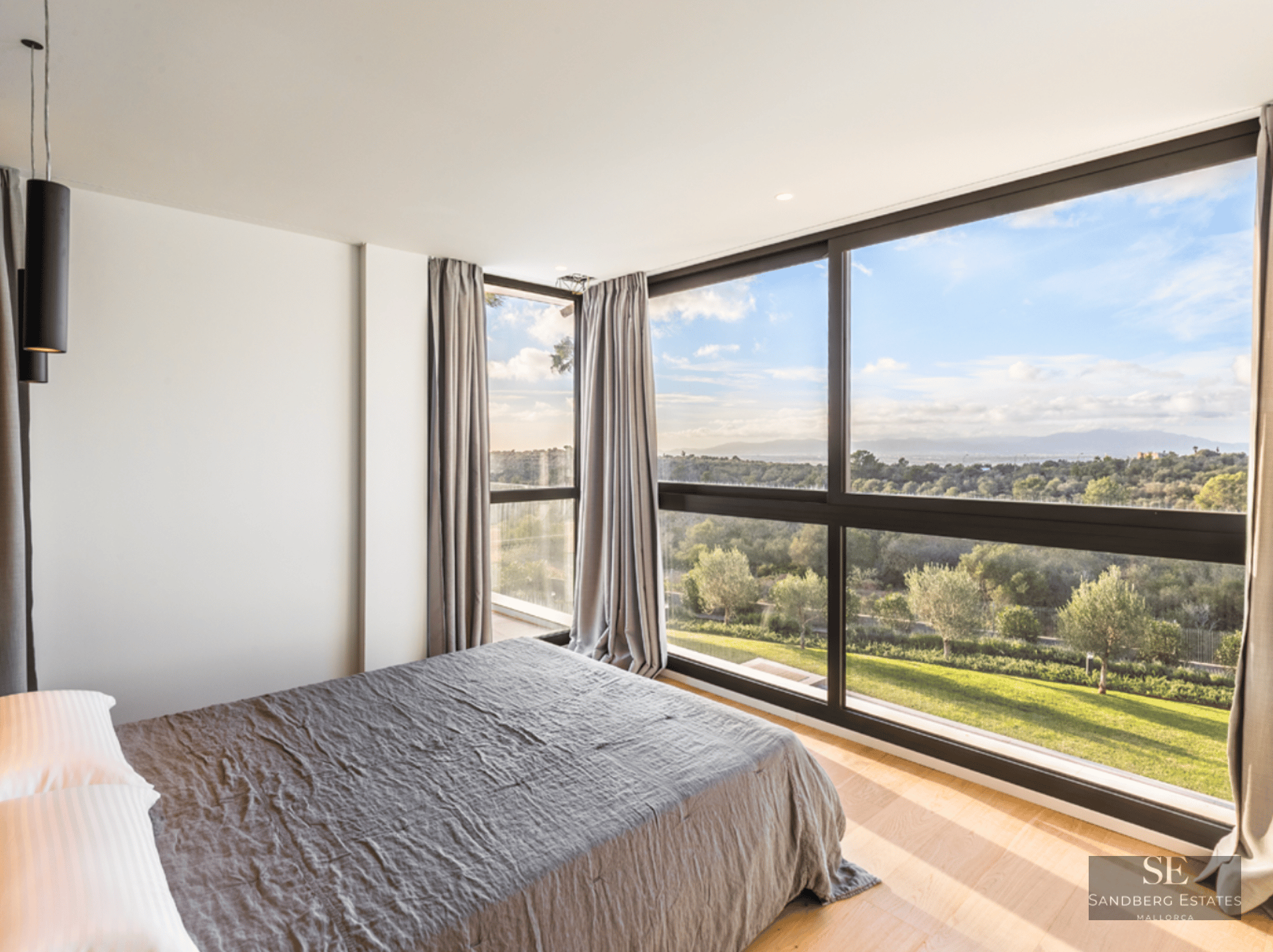 Modern bedroom with large bed and floor-to-ceiling windows overlooking a green landscape and blue sky.